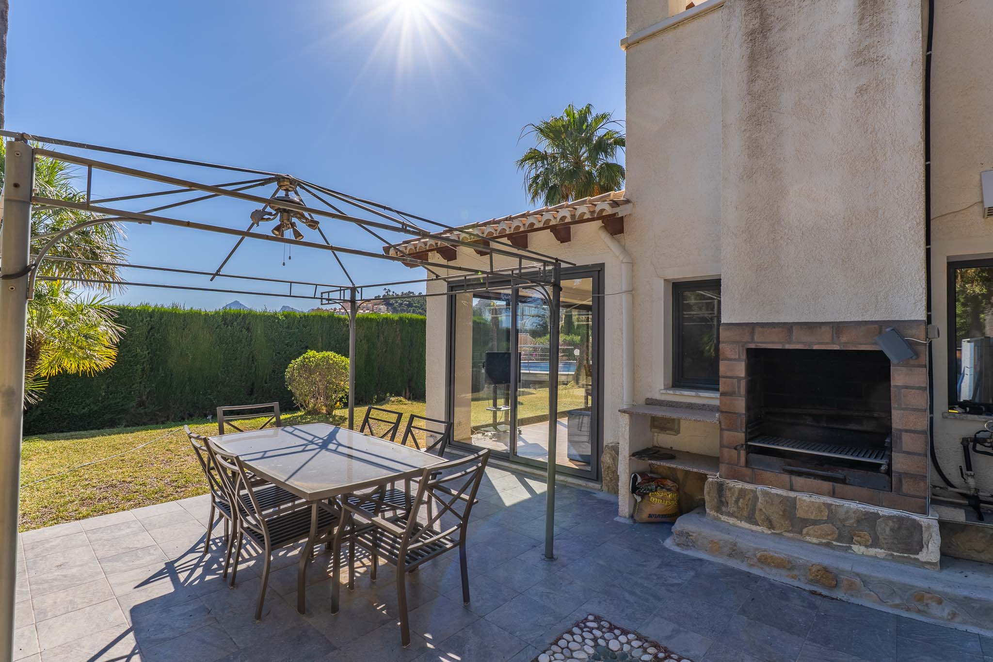 Buitenpatio met donkergrijze tegels, een metalen pergola boven een eettafel en een ingebouwde bakstenen barbecue. Glazen schuifdeuren bieden toegang tot binnen.