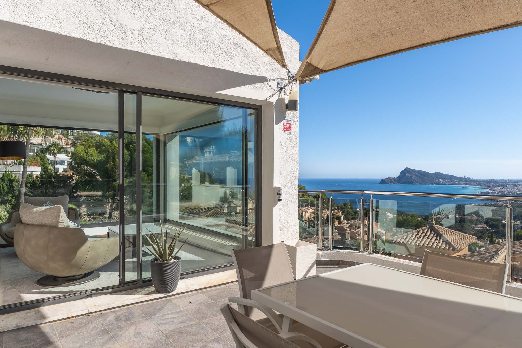 Buitenterras met eettafel en stoelen, deels overschaduwd. Het biedt panoramisch zee- en kustzicht via een glazen balustrade, en verbindt met een lichte binnenruimte.