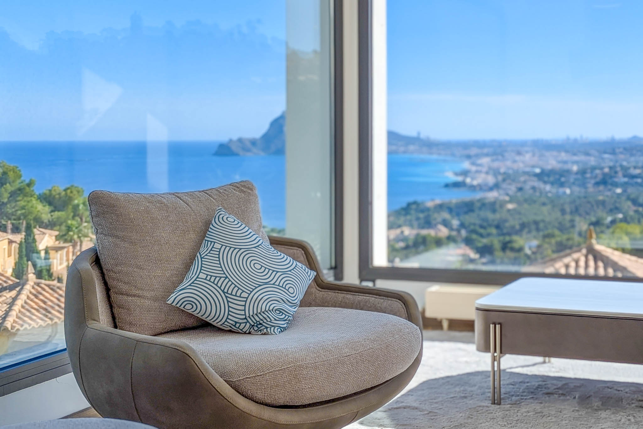 Comfortabele fauteuil bij een panoramisch raam, met ononderbroken uitzicht op de Middellandse Zee en de kustlijn. Een sierkussen voegt een modern accent toe aan het interieur.