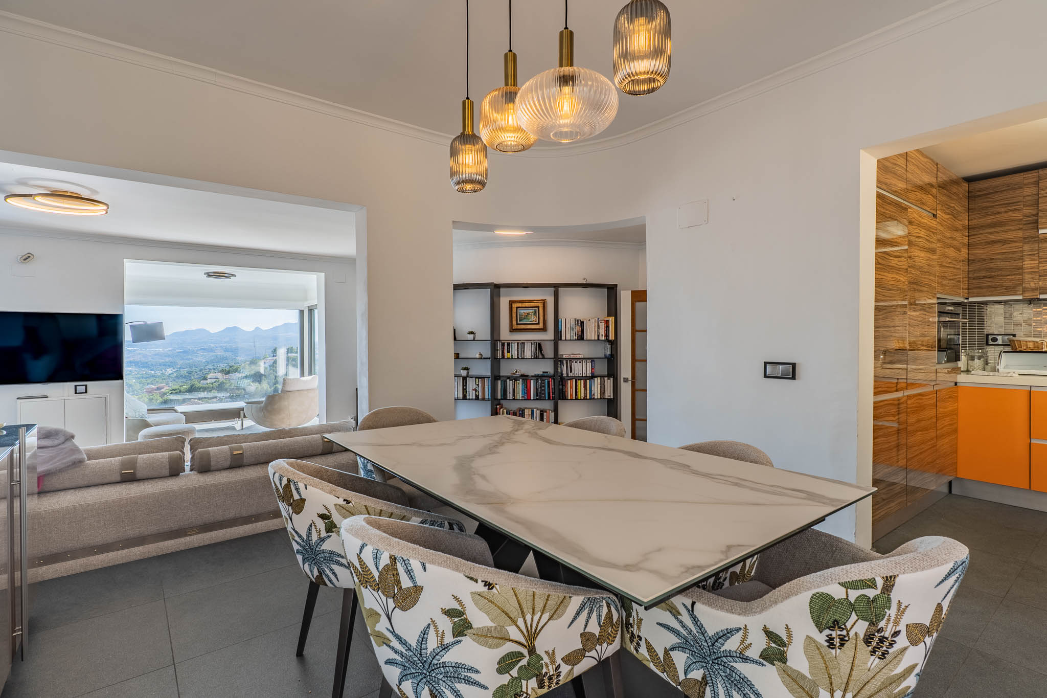 Design eetruimte met marmer-effect tafel en gestoffeerde stoelen, verlicht door moderne hanglampen. Grenst aan de woonkamer met panoramisch uitzicht op de bergen.