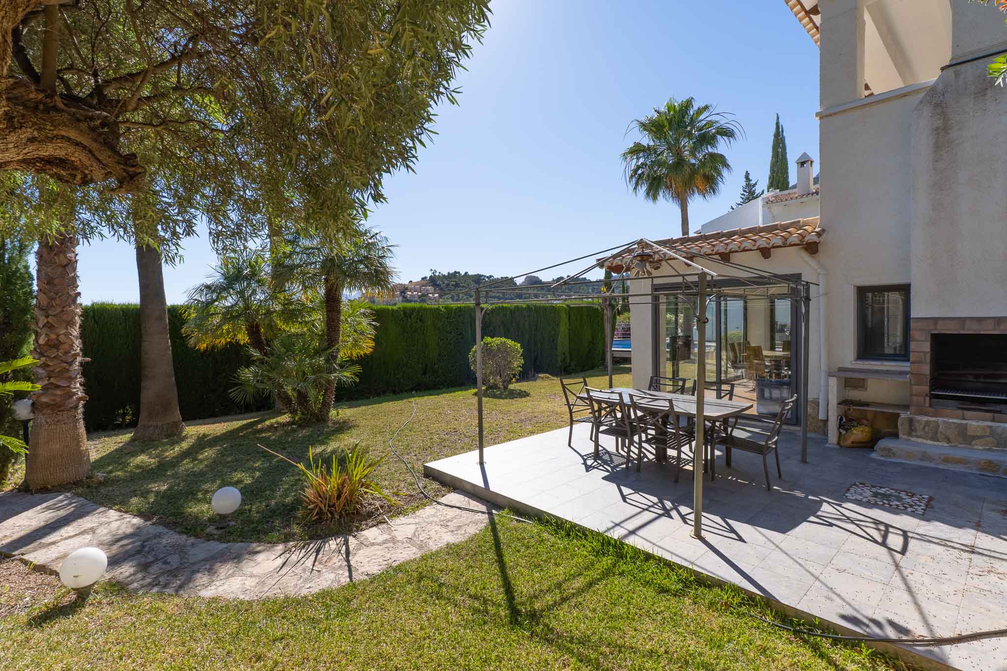 Deze uitnodigende buiten eetruimte omvat een betegeld terras met metalen pergola, ideaal voor al fresco dineren. Het wordt aangevuld met een ingebouwde barbecue en weelderige tuin met palmbomen en haag.