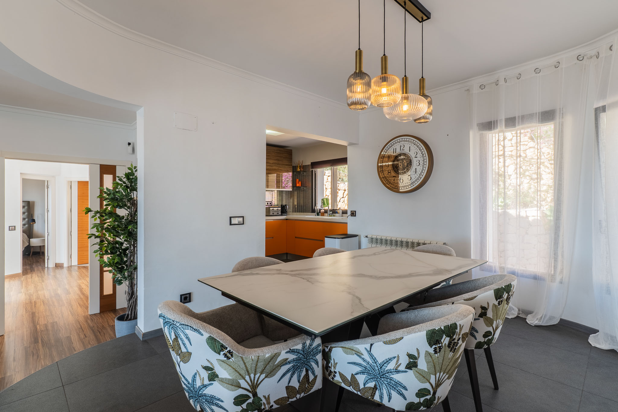 Eetruimte met marmerlook tafel en stoelen met tropisch patroon. Moderne hanglampen verlichten de ruimte, die open is naar een keuken met oranje kasten.