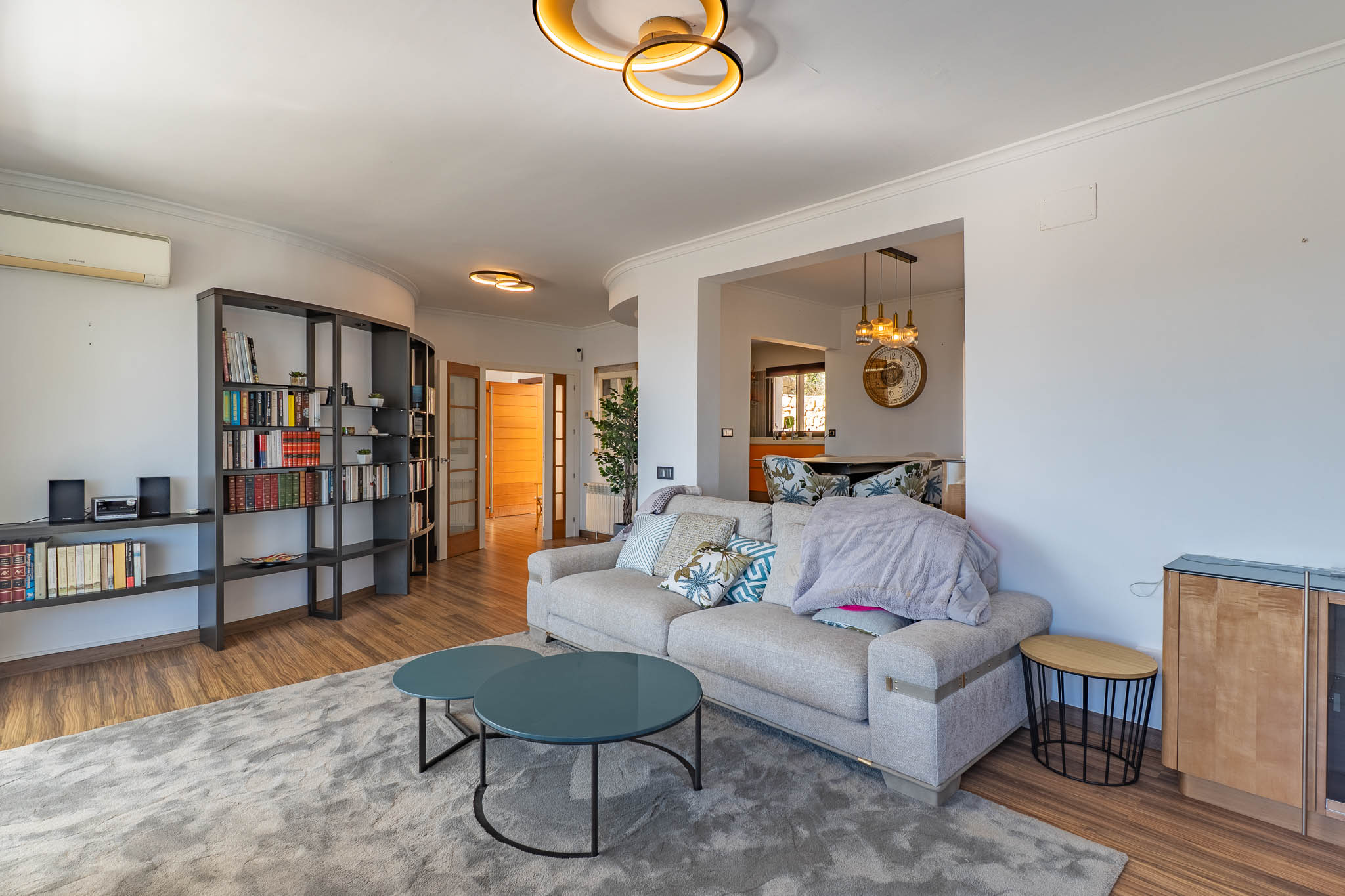 Hedendaagse woonkamer met lichte bank, twee ronde teal salontafels en een grote donkere modulaire boekenkast. Design plafondverlichting siert de ruimte.