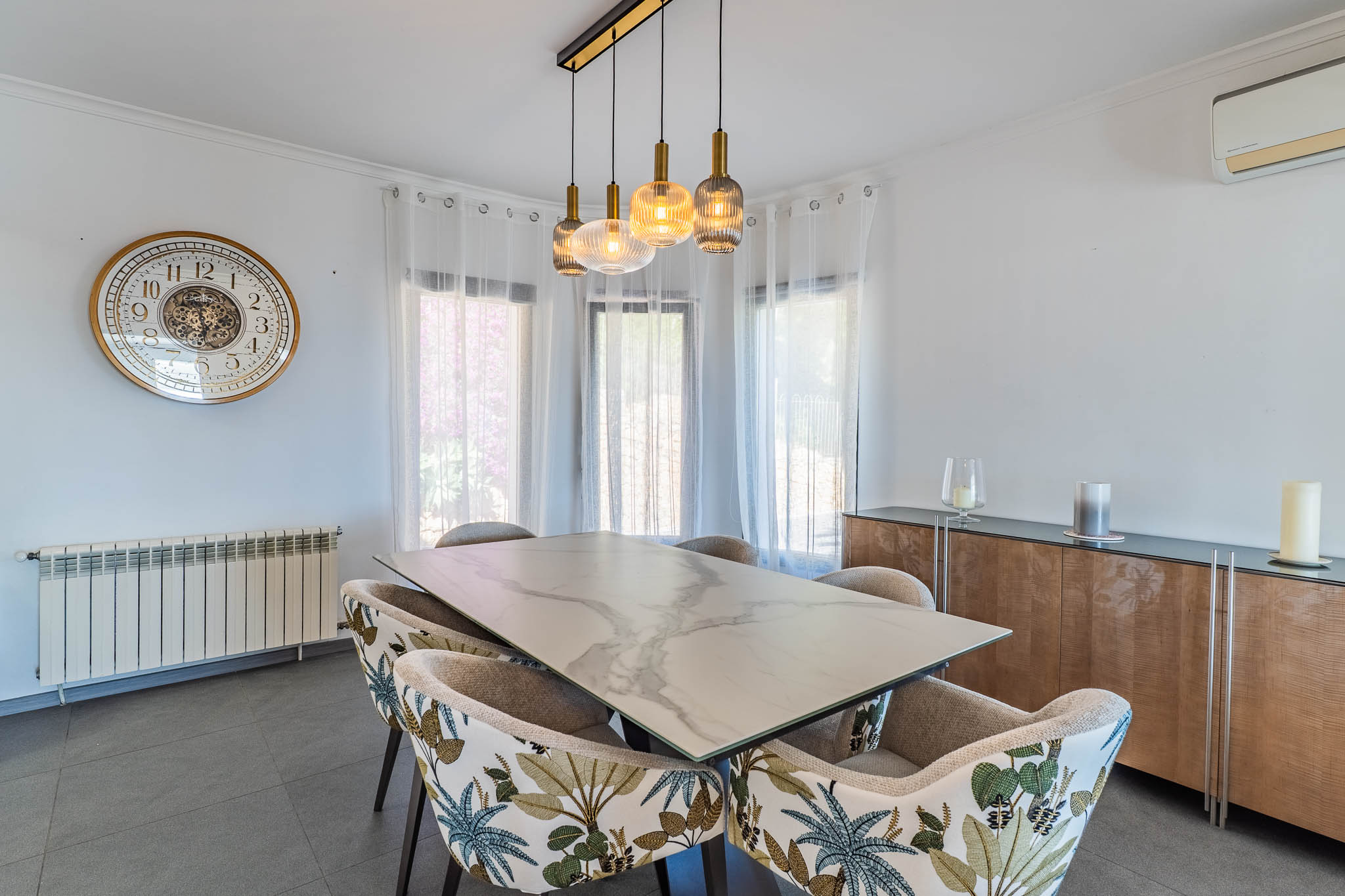 Lichte eetkamer met marmerlook tafel, stoelen met tropisch patroon en een moderne lineaire hanglamp, verlicht door natuurlijk licht.