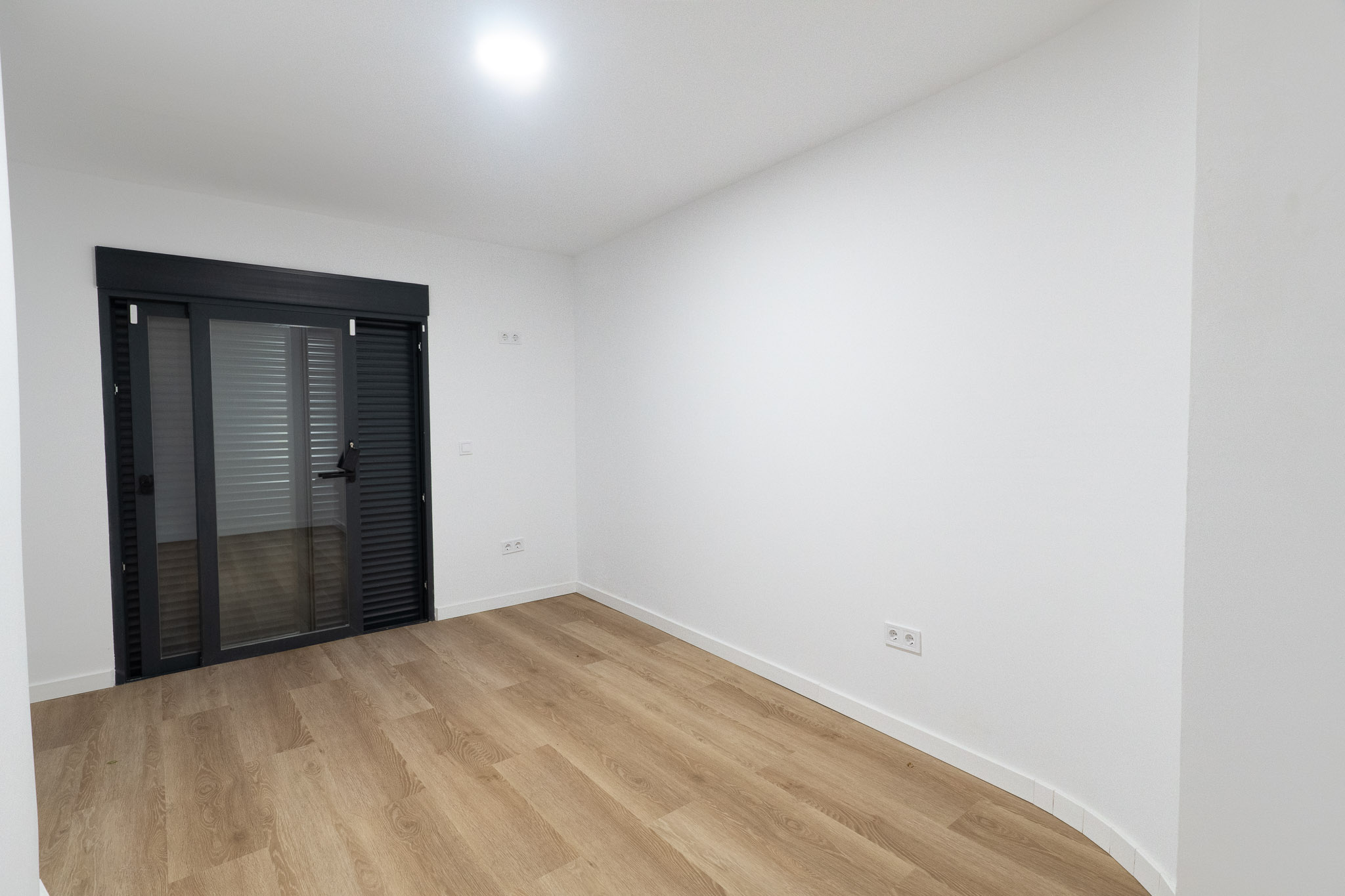 Lichte kamer met lichte houtnerf laminaatvloer en witte muren. Een donkergrijze glazen schuifdeur met geïntegreerde jaloezieën biedt toegang tot buiten.