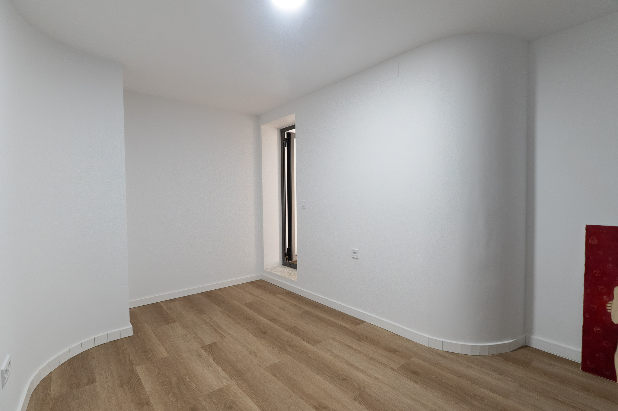 Lichte ruimte met lichte plankenvloer, witte muren en een opvallende gebogen wand. Voorzien van een smalle donker omlijste opening met stenen dorpel.