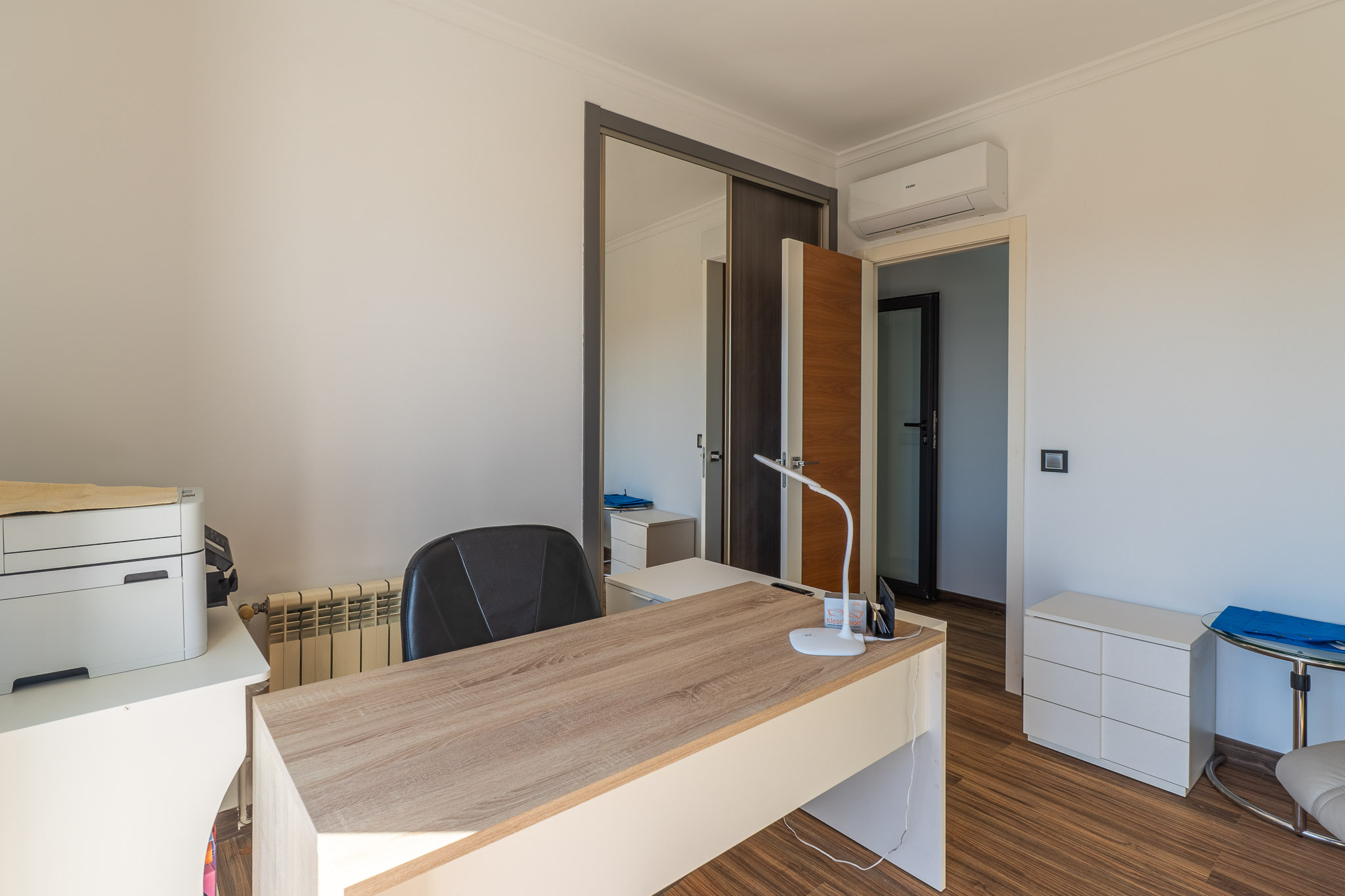 Lichte studeerkamer met licht houten bureau, laminaatvloer, spiegelende schuifdeurkast en een airconditioningunit.