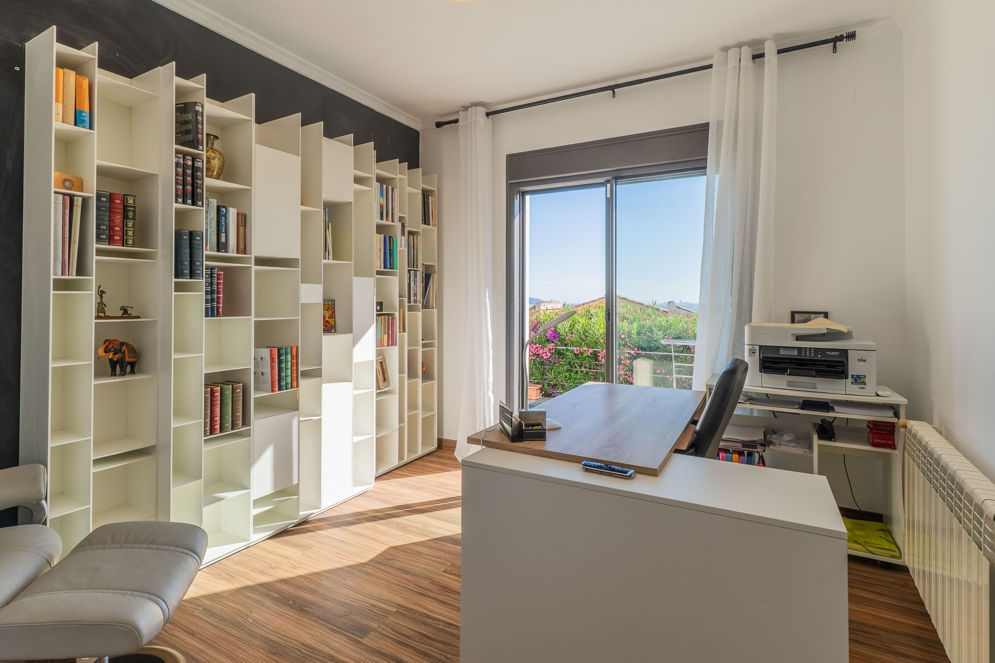 Lichte thuiskantoor met een wit modulair boekenkastsysteem, houten vloer en een modern bureau voor een balkon met weelderig groen uitzicht. Natuurlijk licht vult de ruimte.