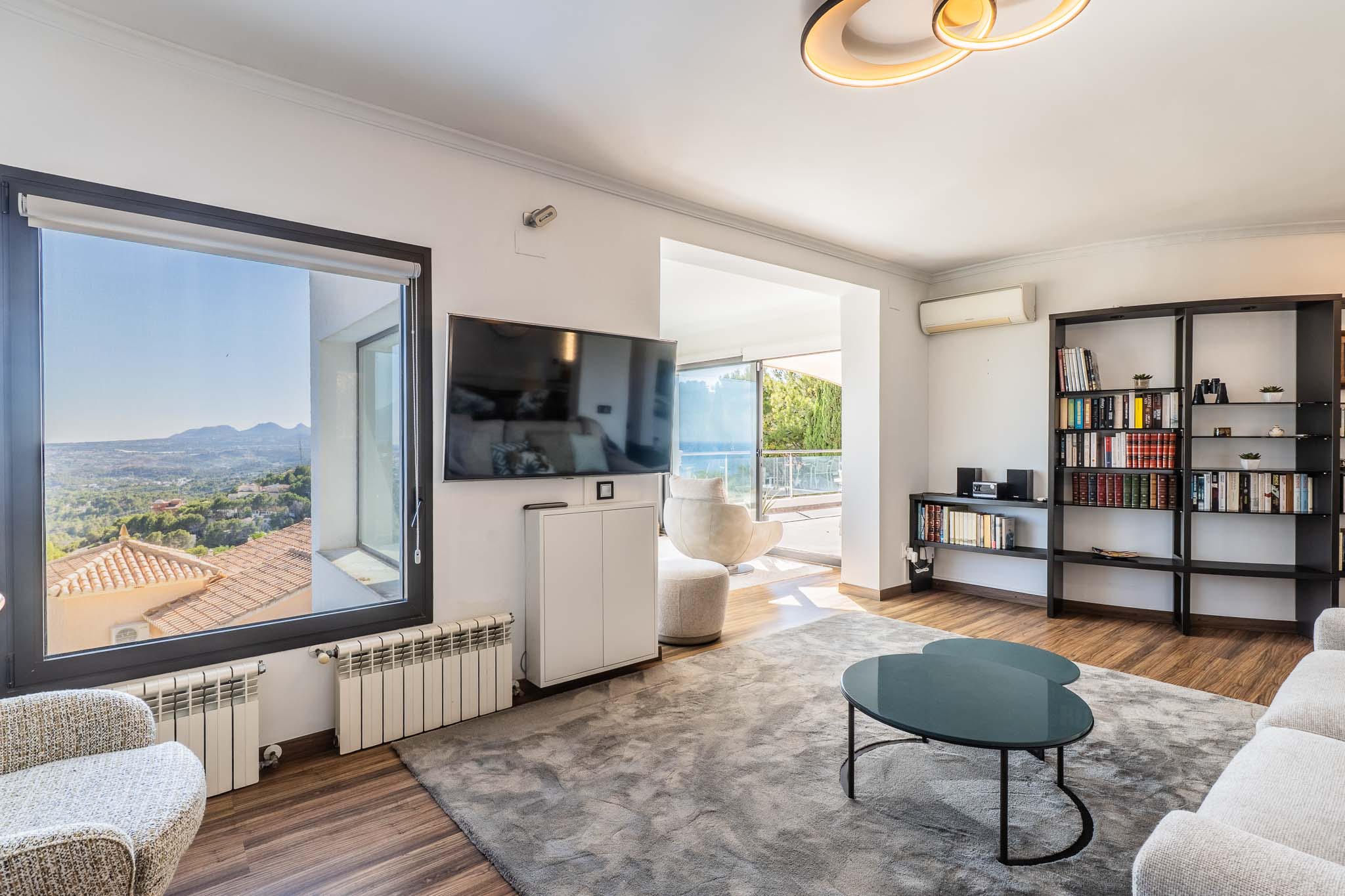Lichte woonkamer met panoramisch uitzicht op de bergen vanuit het raam en toegang tot het terras met zeezicht. Voorzien van een houten vloer, een groot grijs vloerkleed en een moderne boekenkast.