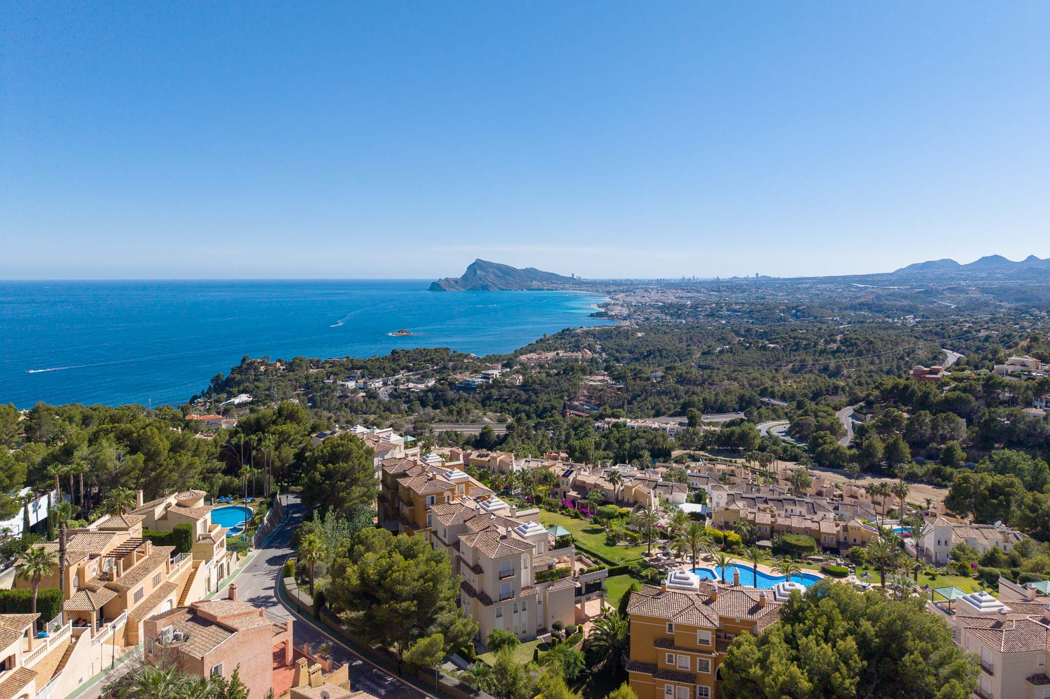 Luchtfoto van de kustlijn van Altea Hills, met de Middellandse Zee, de Peñón de Ifach en de skyline van Benidorm in de verte. Terracotta daken en zwembaden zijn zichtbaar tussen weelderig groen.