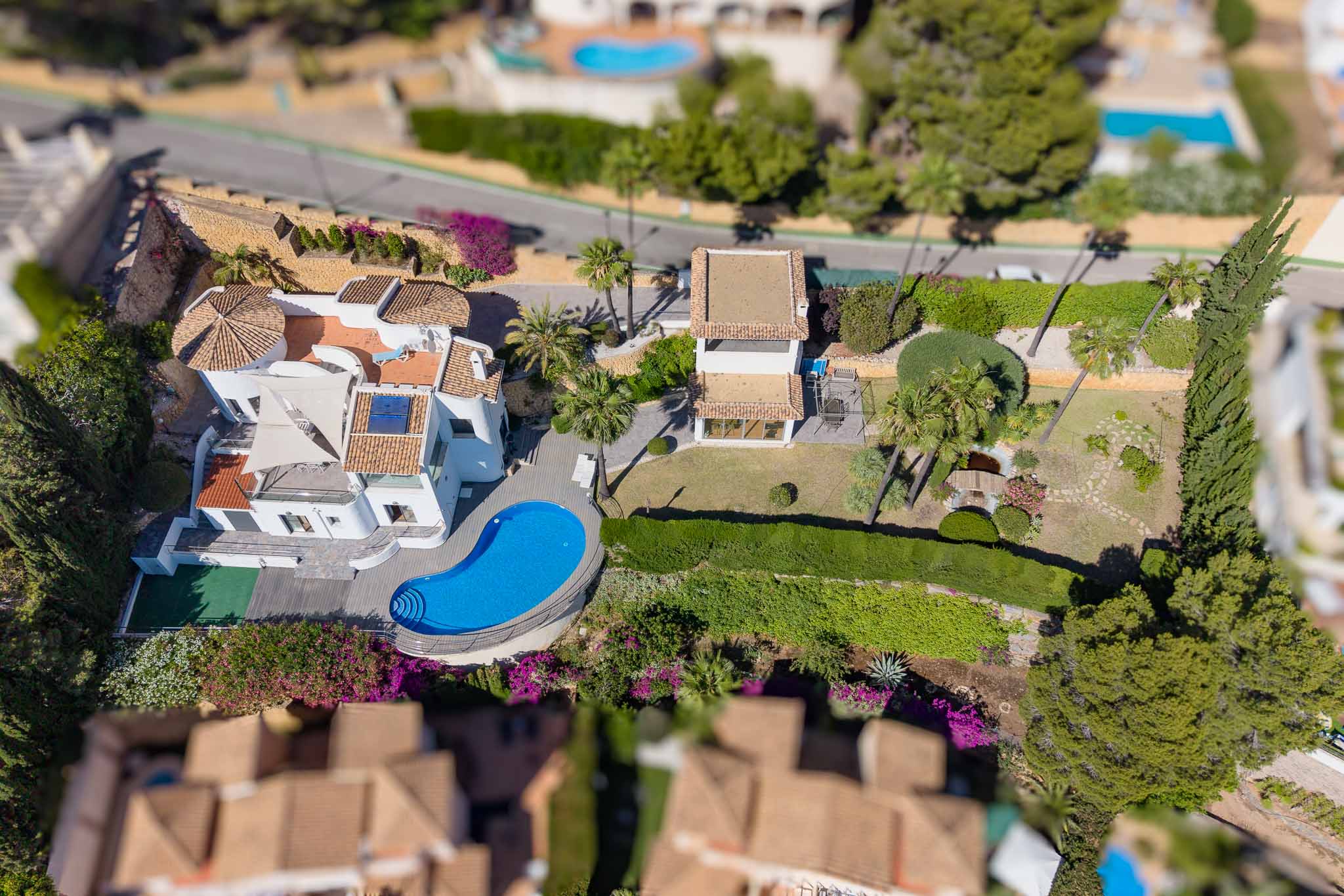 Luchtfoto van de villa met onregelmatig gevormd zwembad, uitgestrekte vlakke tuinen met vijver en een apart bijgebouw. Opvallend zijn de traditionele pannendaken en weelderige mediterrane beplanting.