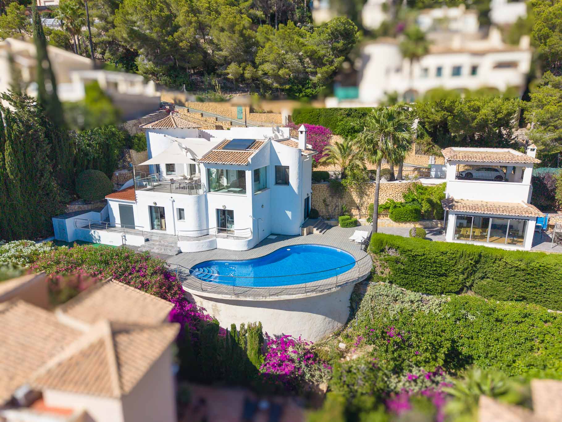 Luchtfoto van de witte villa, met een uniek niervormig zwembad omringd door een houten terras en metalen reling. Meerdere terrassen en weelderige mediterrane tuinen zijn zichtbaar.