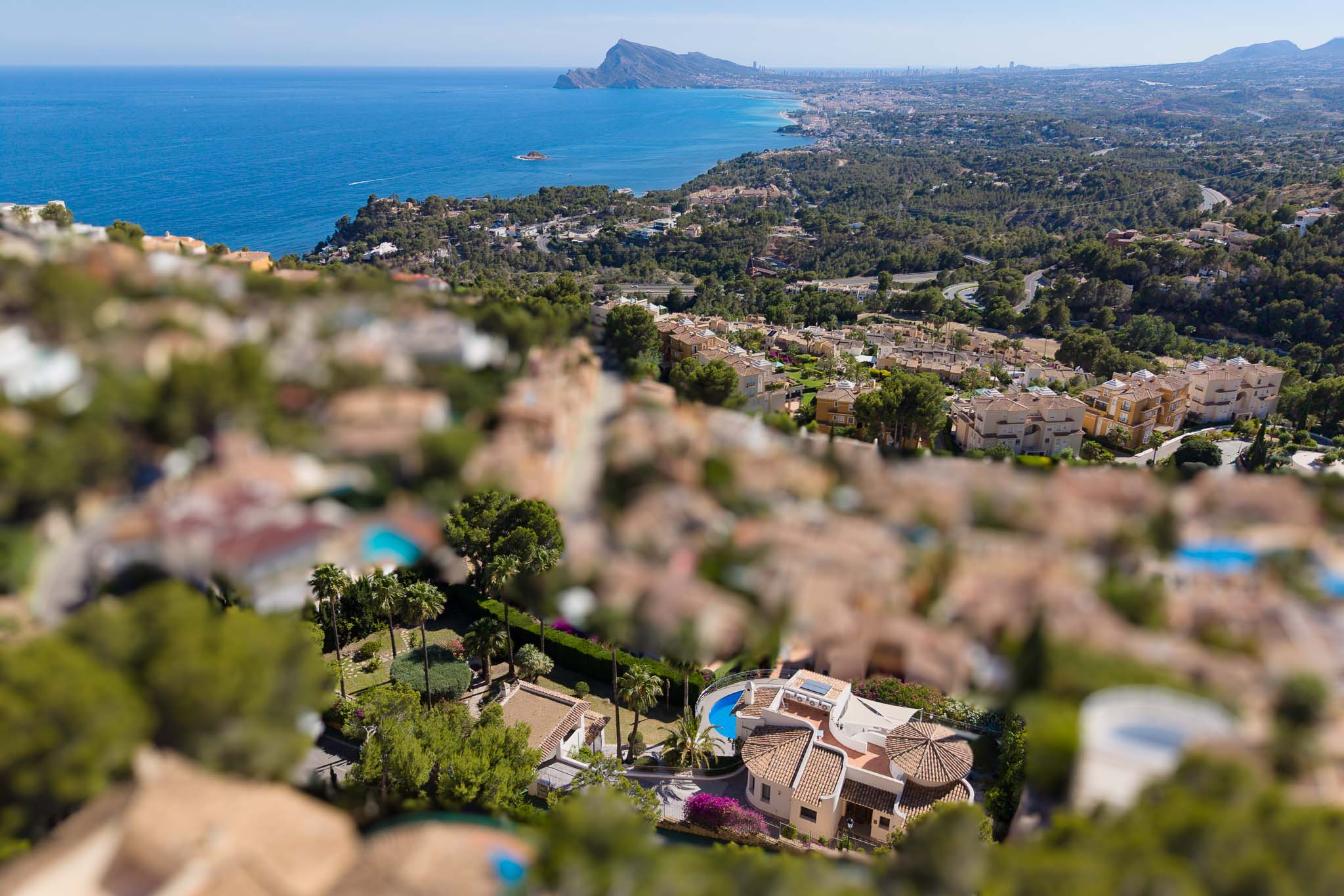 Luchtfoto van een villa in Altea Hills met een gebogen zwembad, terracotta daken en weelderige tuinen, met de Middellandse Zee en kustlijn op de achtergrond.