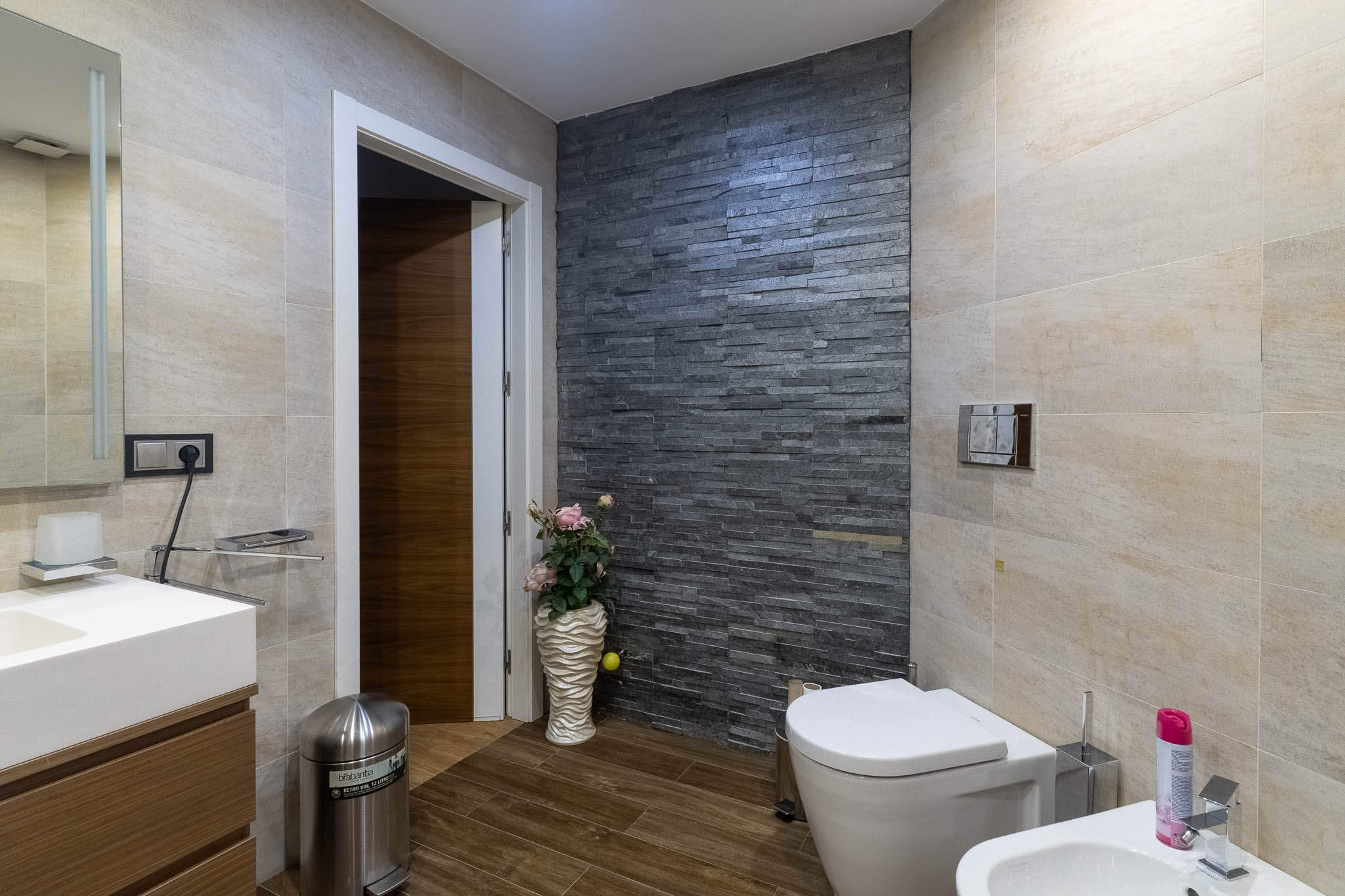 Moderne badkamer met donkere gestapelde stenen accentmuur, lichtbeige tegels en houtlookvloer. Voorzien van toilet, bidet en badmeubel met wastafel.