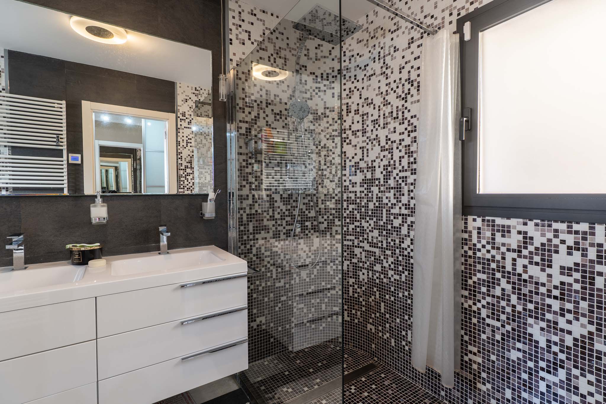 Moderne badkamer met dubbele wastafel, wit meubel en verlichte spiegel. De glazen douchewand heeft mozaïektegels in grijstinten en een geïntegreerd raam.
