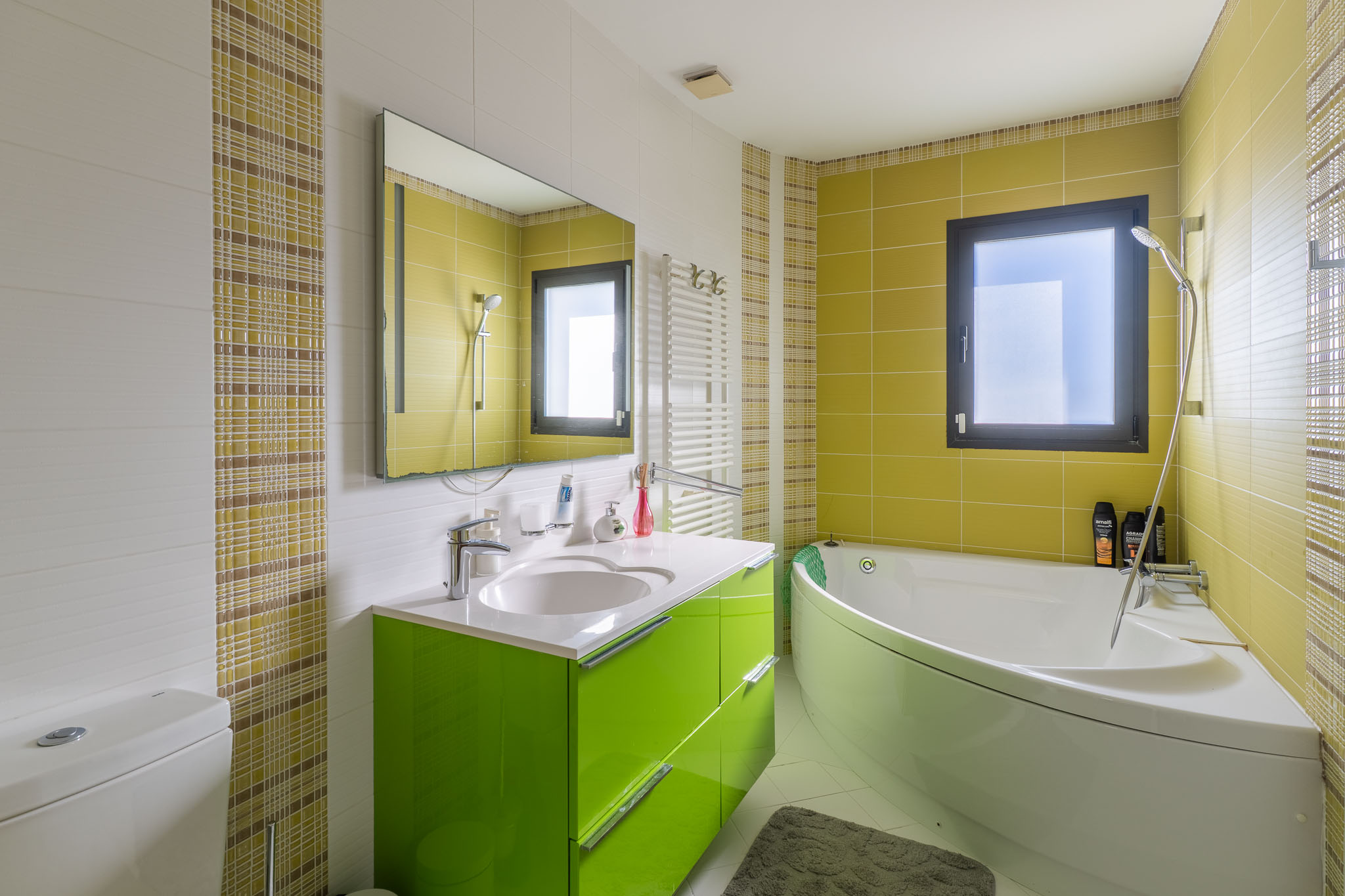 Moderne badkamer met hoekbad en een felgroen wastafelmeubel. Muren voorzien van witte structuur-, limoengroene en geruite mozaïektegels. Inclusief raam en handdoekradiator.