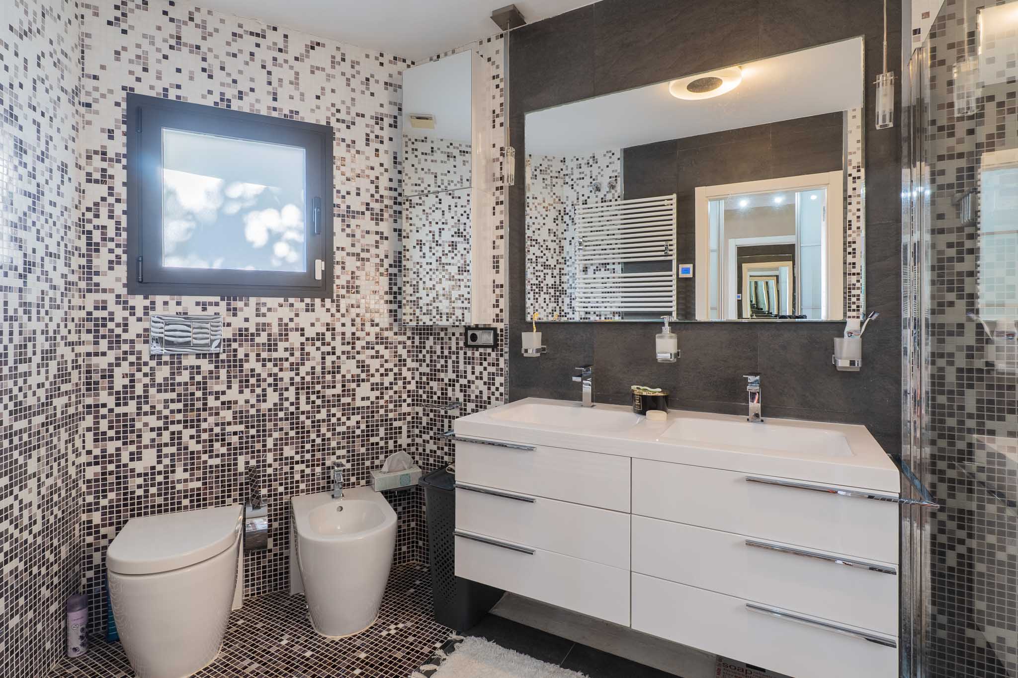 Moderne badkamer met mozaïektegels op wanden en vloer in aardse/grijze tinten. Beschikt over een dubbele zwevende wastafel, grote verlichte spiegel, wandtoilet, bidet en handdoekradiator.