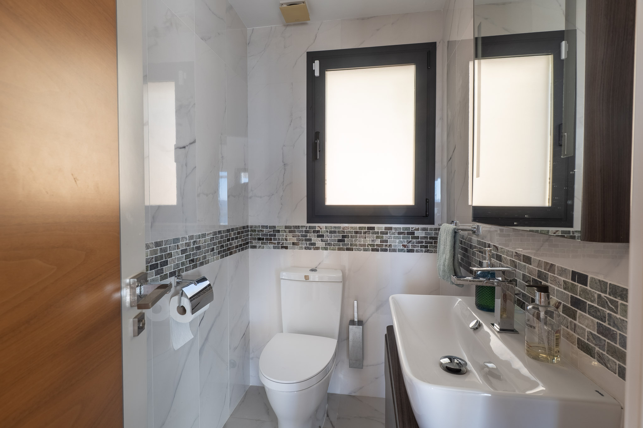Moderne badkamer met witte marmerlook tegels, een grijze mozaïekrand en een donker kozijnraam. Voorzien van een strakke witte wastafel en bijpassend toilet.