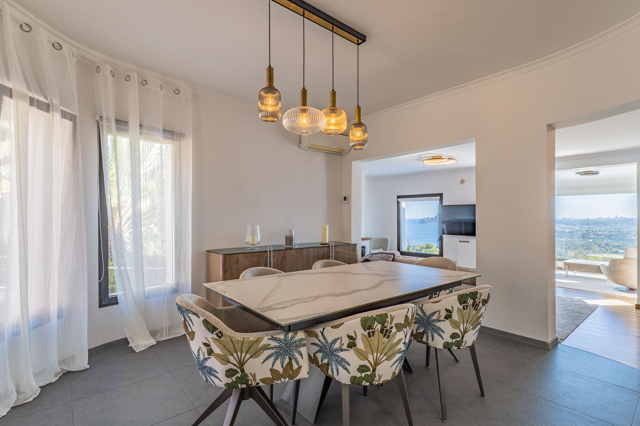 Moderne eetkamer met marmerlook tafel en stoelen met tropisch bladpatroon. Een lineaire kroonluchter verlicht de ruimte, die vanuit aangrenzende kamers panoramisch uitzicht op zee biedt.