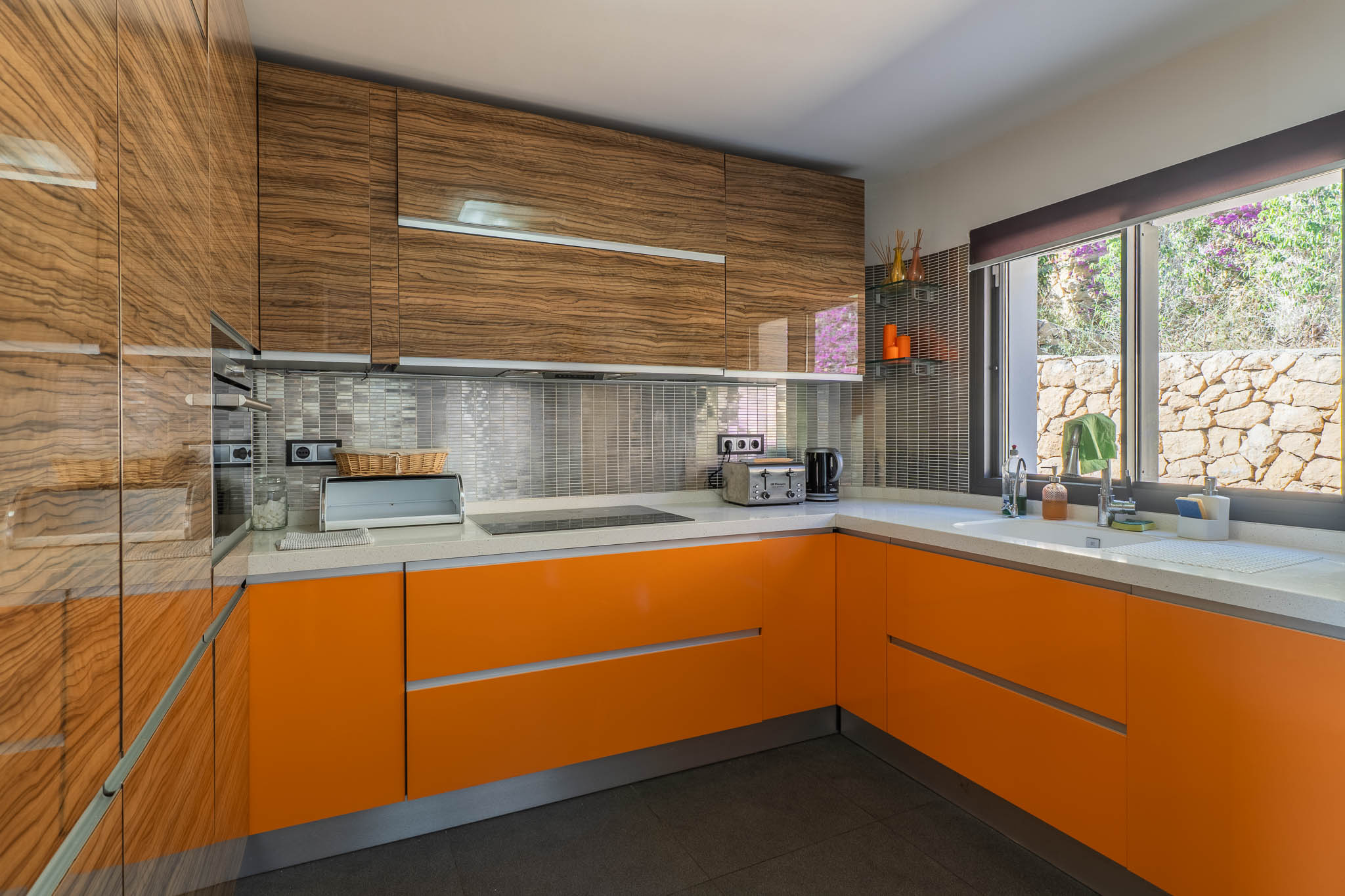 Moderne keuken met glanzende bovenkasten in houtnerf en feloranje onderkasten. Wit aanrechtblad, metallic mozaïek achterwand en een raam met uitzicht op een stenen muur.