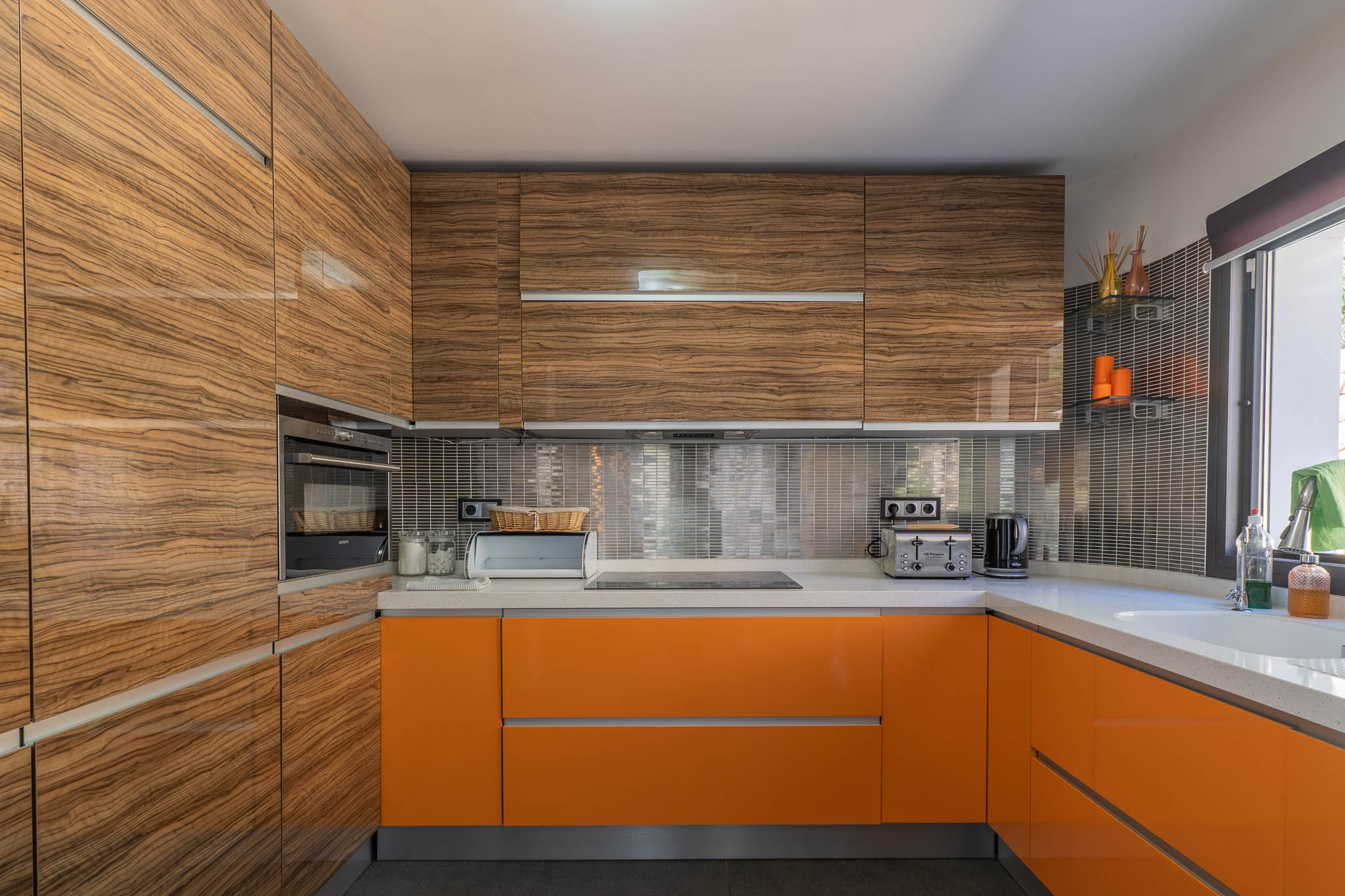 Moderne keuken met glanzende houtnerf bovenkasten en levendige oranje onderkasten, aangevuld met lichte werkbladen en een metallic mozaïek achterwand.