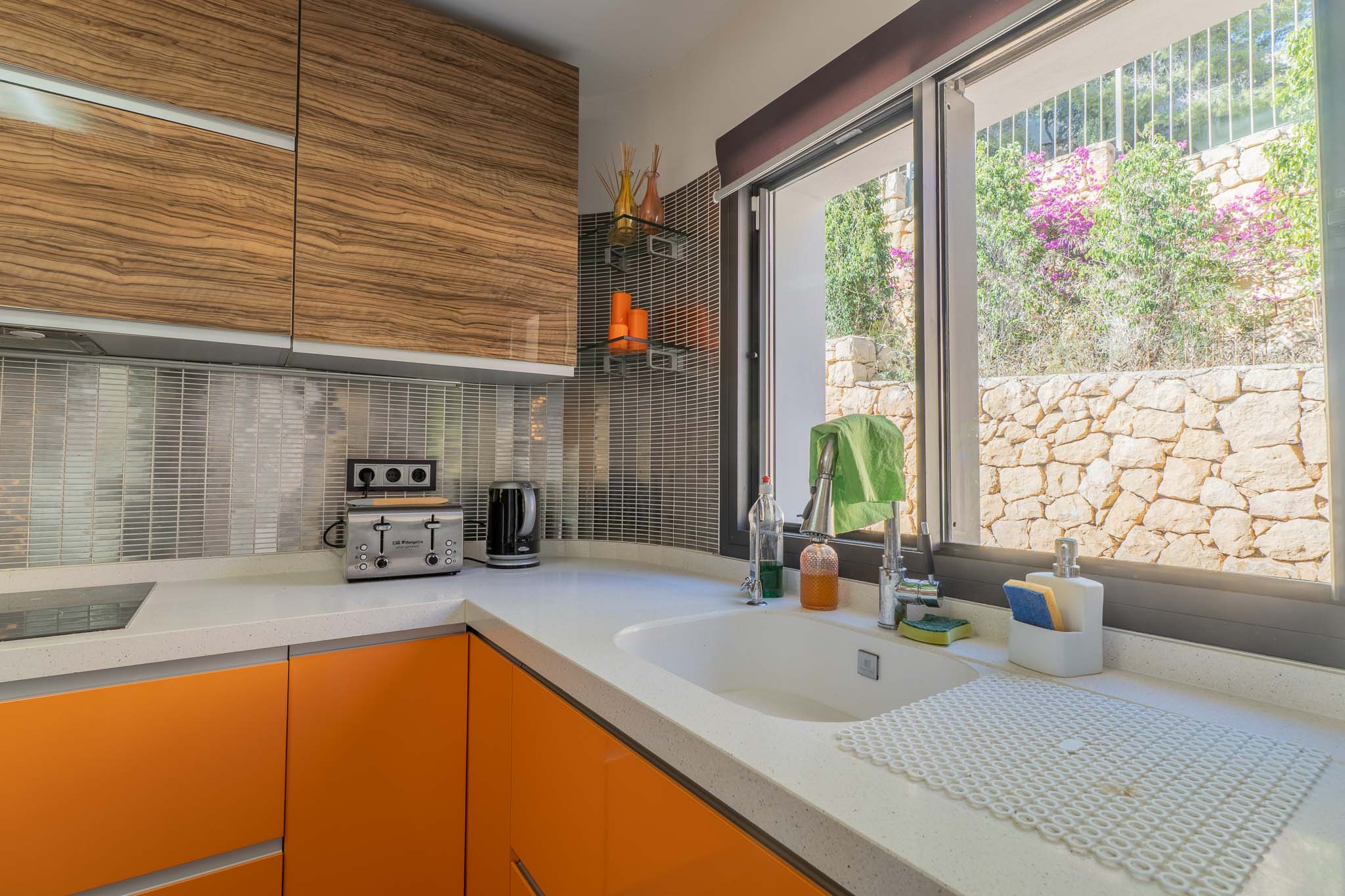 Moderne keuken met wit kwartswerkblad, oranje onderkasten en bovenkasten met houtnerf. Het raam biedt uitzicht op een natuurstenen muur en weelderig groen.