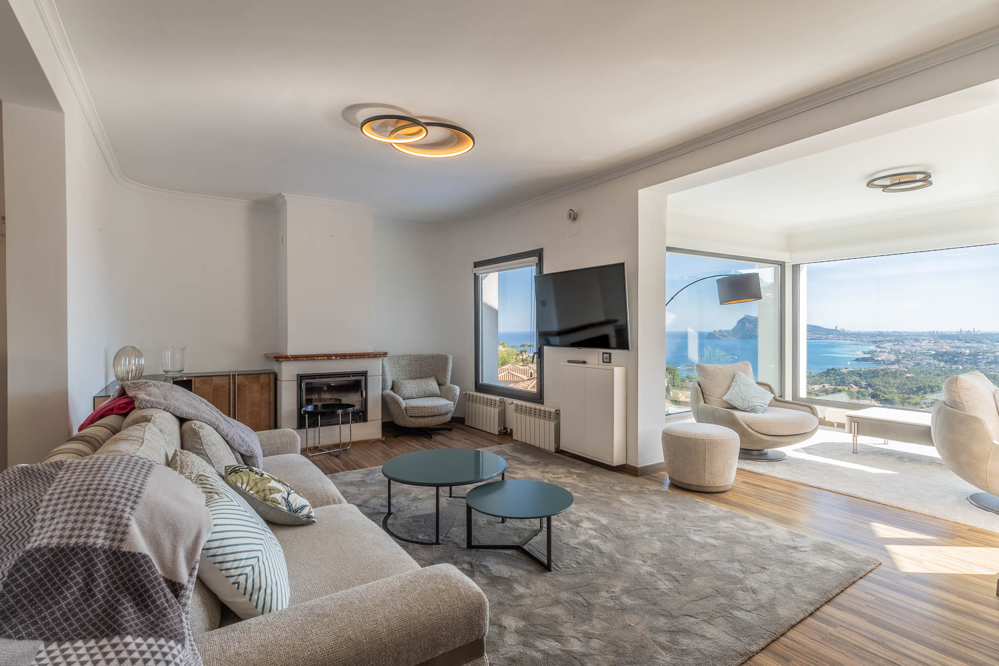 Moderne woonkamer met lichte bank, open haard en grote ramen die een panoramisch uitzicht op zee en stad bieden. Hedendaags meubilair versterkt de lichte ruimte.