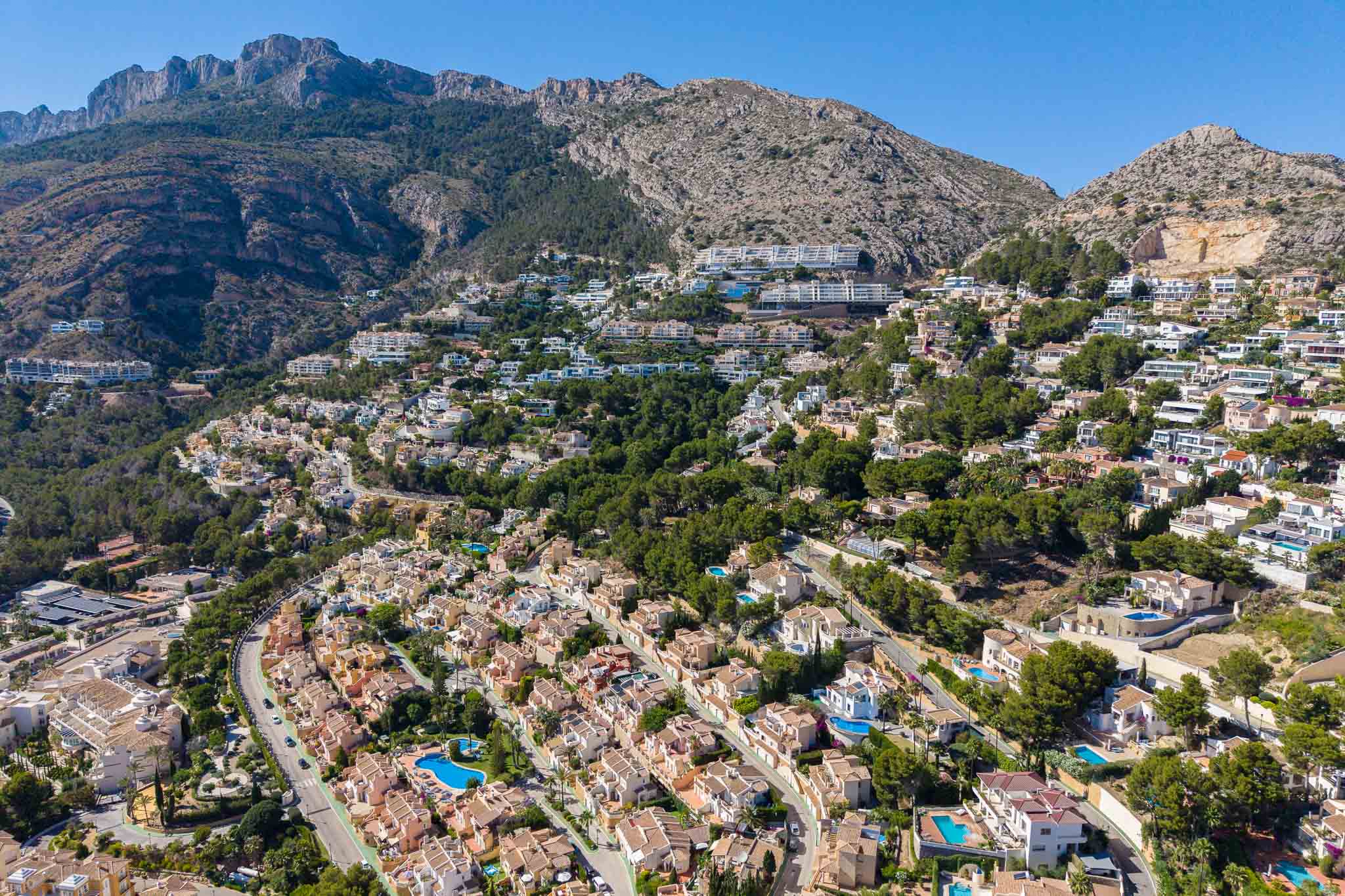 Panoramisch luchtbeeld van Altea Hills, met talloze villa's met zwembaden op de groene heuvel, tegen een indrukwekkend berglandschap.