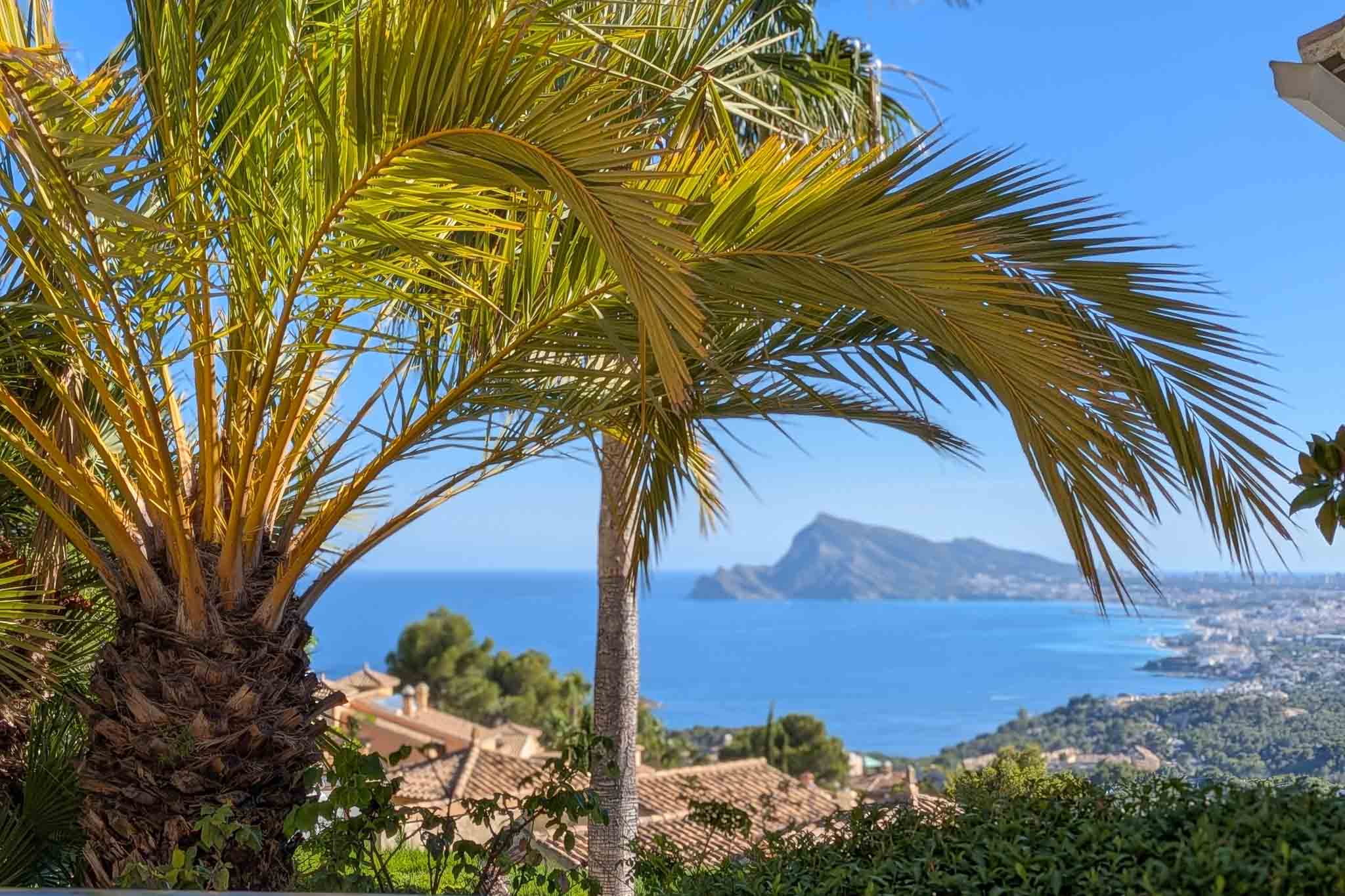 Panoramisch uitzicht op de Middellandse Zee, omlijst door palmbomen, met de daken van Altea Hills en de verre Peñón de Ifach onder een helderblauwe lucht.