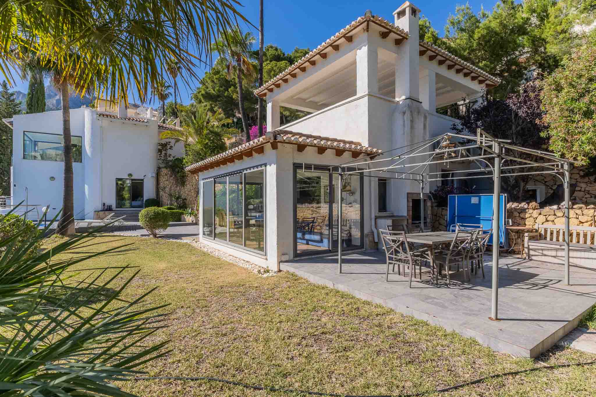 Ruim betegeld terras met eethoek onder een metalen pergola, grenzend aan een glazen serre en een verzorgde mediterrane tuin.