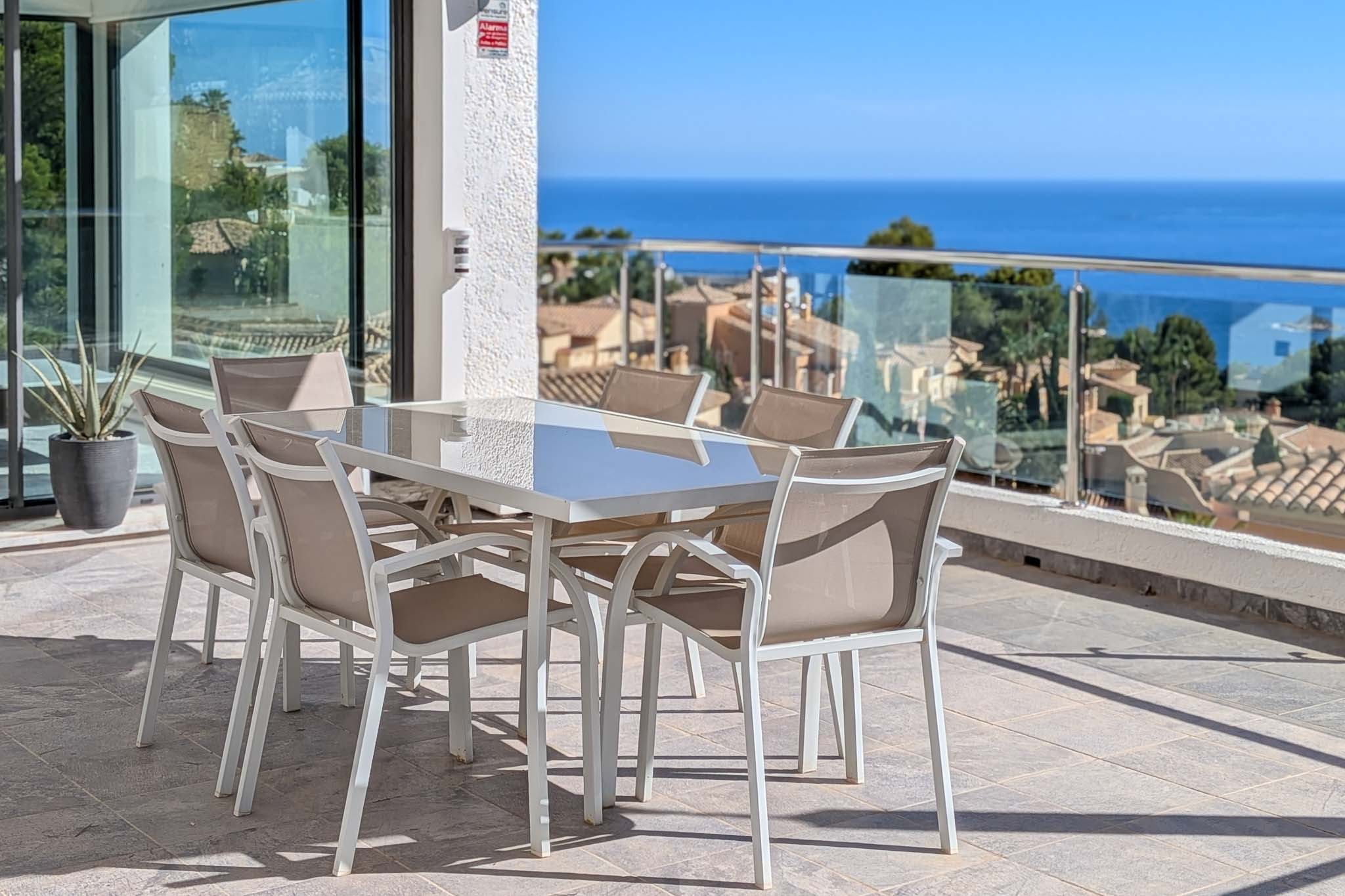 Ruim buitenterras met eettafel en stoelen, met panoramisch uitzicht op de Middellandse Zee en de daken van Altea Hills. Glazen schuifdeuren verbinden met het interieur.