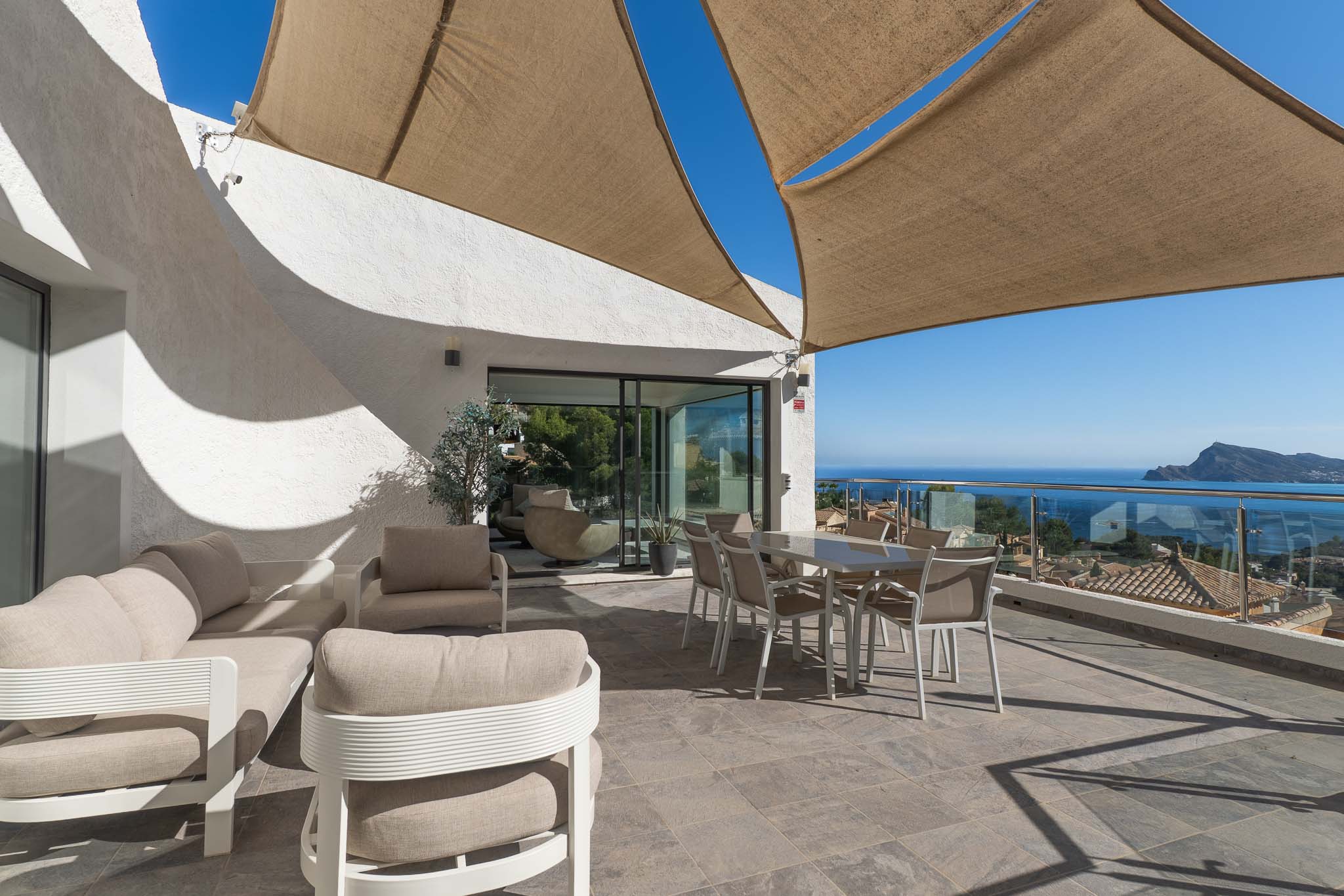 Ruim buitenterras met lounge- en eethoek onder een schaduwdoek, met panoramisch zee- en kustzicht via een glazen balustrade.