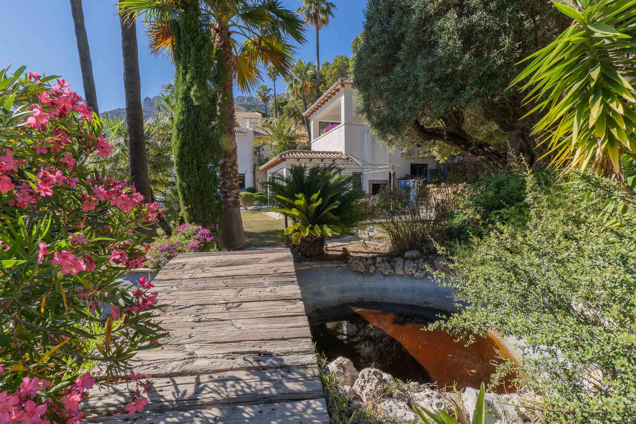 Rustieke houten loopbrug over een serene vijver, omzoomd door levendige roze oleanders en hoge palmbomen, leidend naar de villa onder de mediterrane zon.