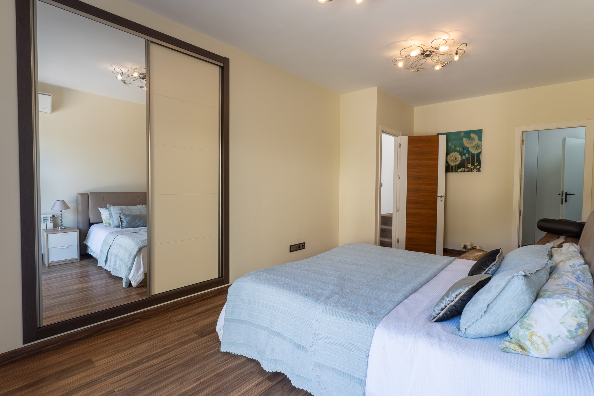 Slaapkamer met bed in lichtblauwe stoffen, houten laminaatvloer en schuifkast met spiegelpaneel. Een moderne plafondlamp en bloemenkunst aan de muur zijn aanwezig.