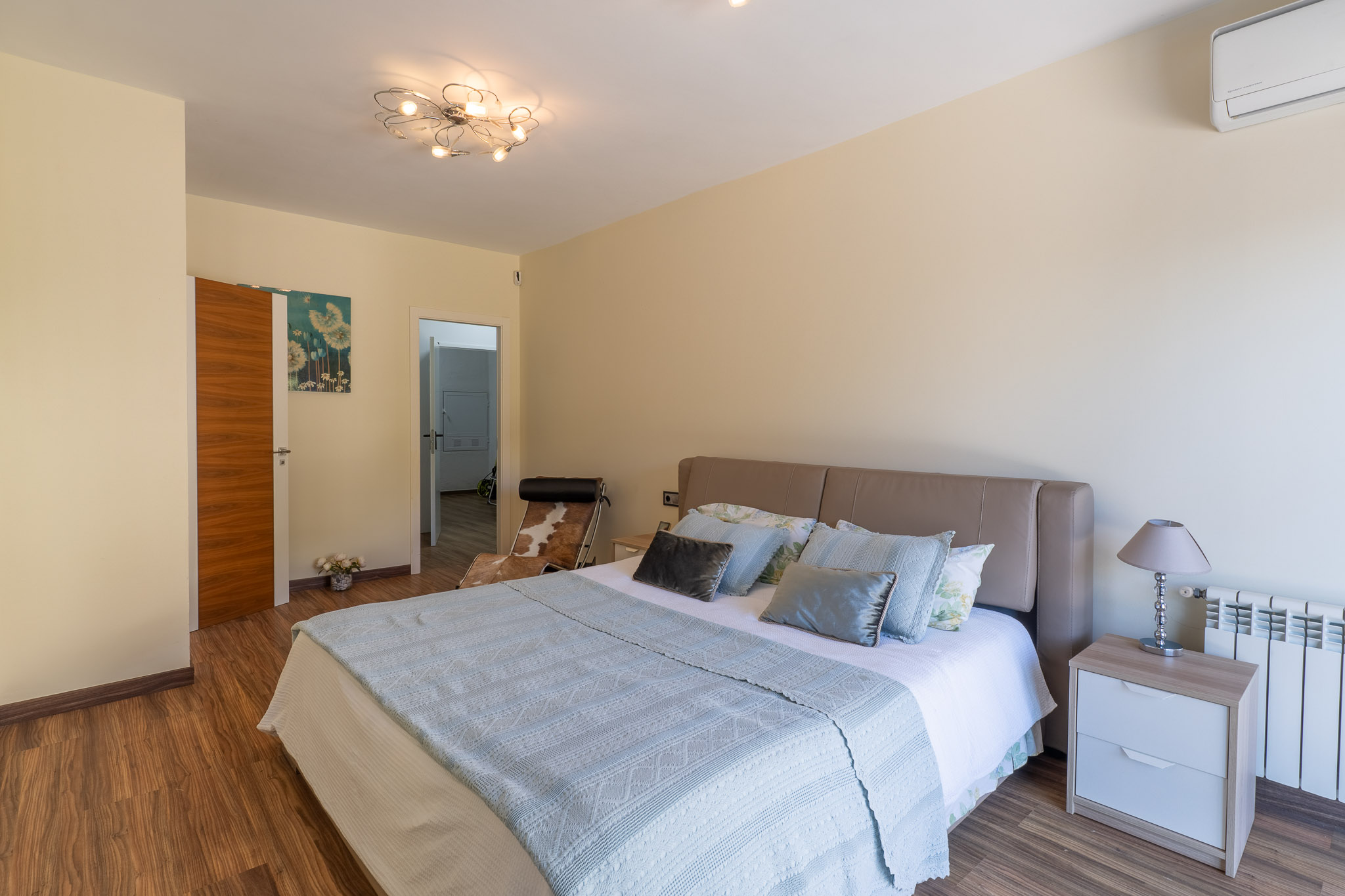 Slaapkamer met beige hoofdeinde, laminaatvloer en moderne plafondlamp. Een nachtkastje met lamp, radiator en airconditioning zijn zichtbaar.