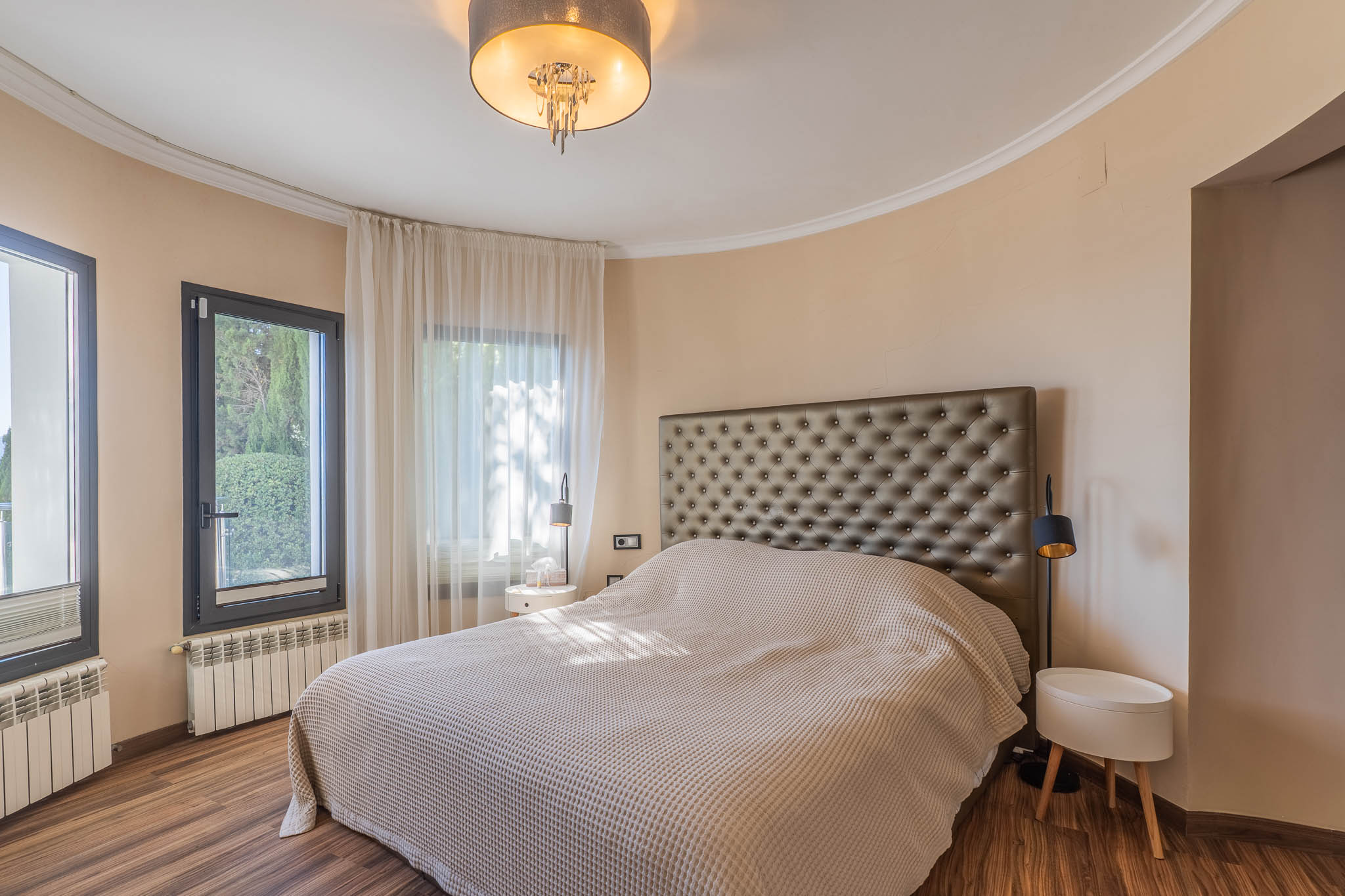 Slaapkamer met groot bed en gecapitonneerd hoofdbord. Gebogen wand met donkeromlijste ramen en lichte gordijnen. Houten vloer, radiatoren en een moderne plafondlamp.