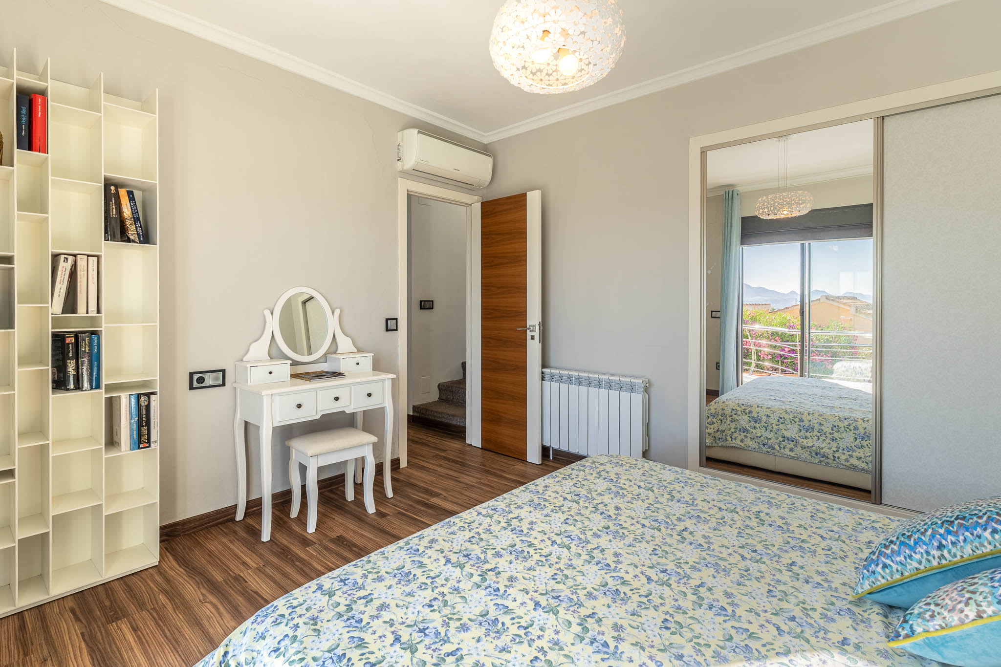 Slaapkamer met laminaatvloer, witte kaptafel en modulair boekenrek. De spiegelkast reflecteert een berguitzicht vanaf het balkon.