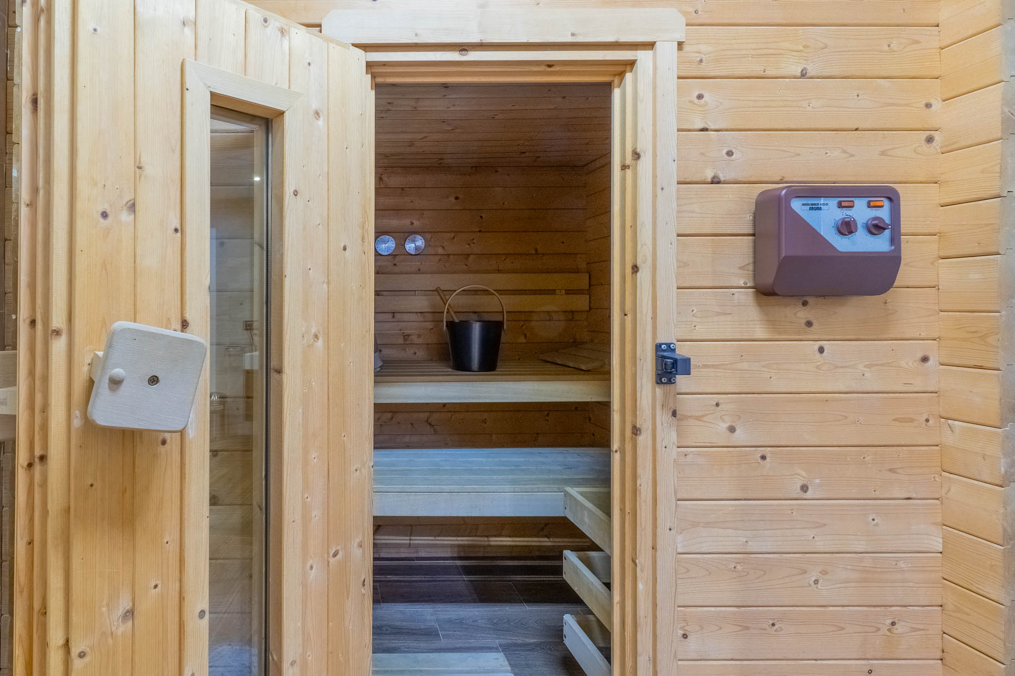 Toegang tot de sauna met lichte houten lambrisering, tonend de binnenbanken, een emmer met lepel en het externe bedieningspaneel.