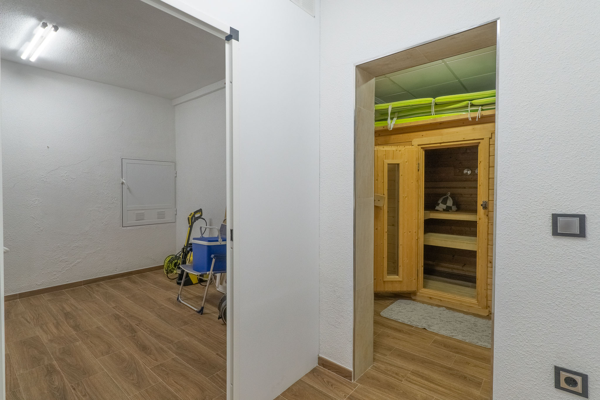 Toegewijde wellnessruimte met houten saunacabine en banken, grenzend aan een praktische bijkeuken. Beide ruimtes hebben een consistente houtnerf tegelvloer.