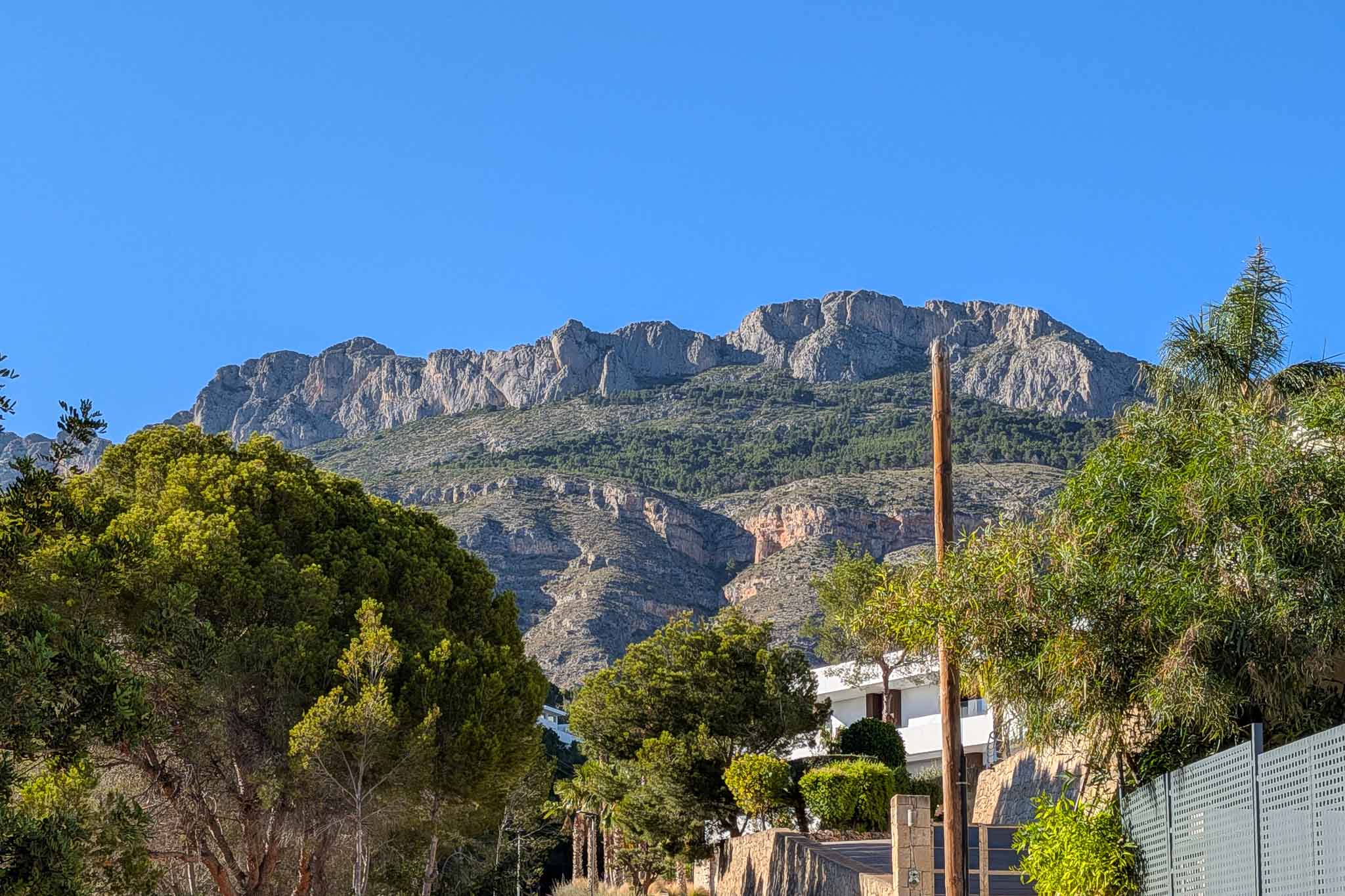 Uitzicht op de majestueuze Sierra de Bernia, met zijn rotsachtige toppen en groene hellingen, omlijst door de weelderige mediterrane vegetatie van het pand.