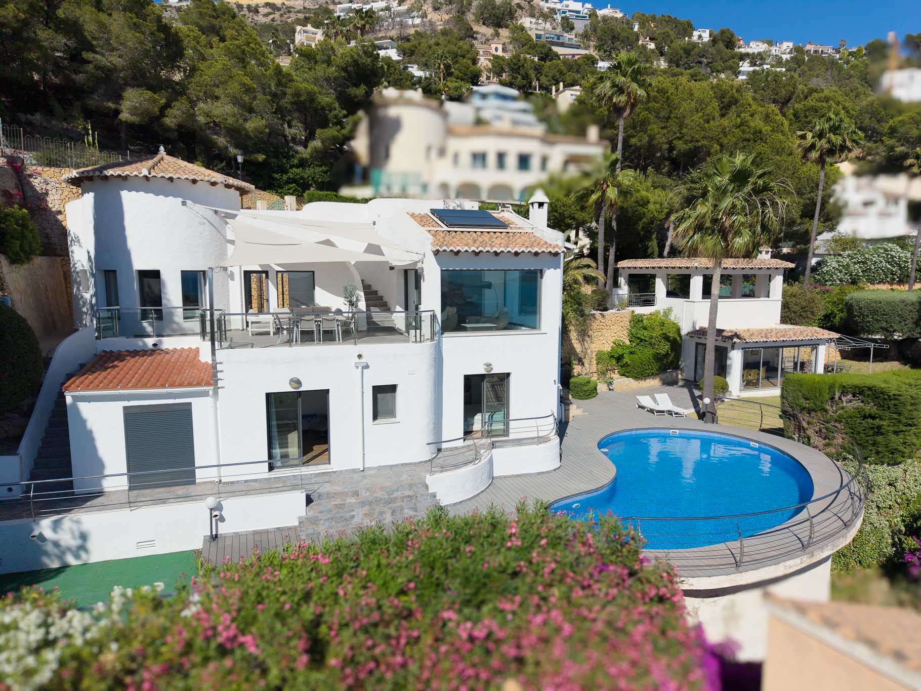 Verhoogd zicht op de hoofdvilla, met een groot boventerras met eethoek en zonnescherm, een niervormig zwembad met terras en een apart gastenverblijf, omgeven door weelderig mediterraan groen.