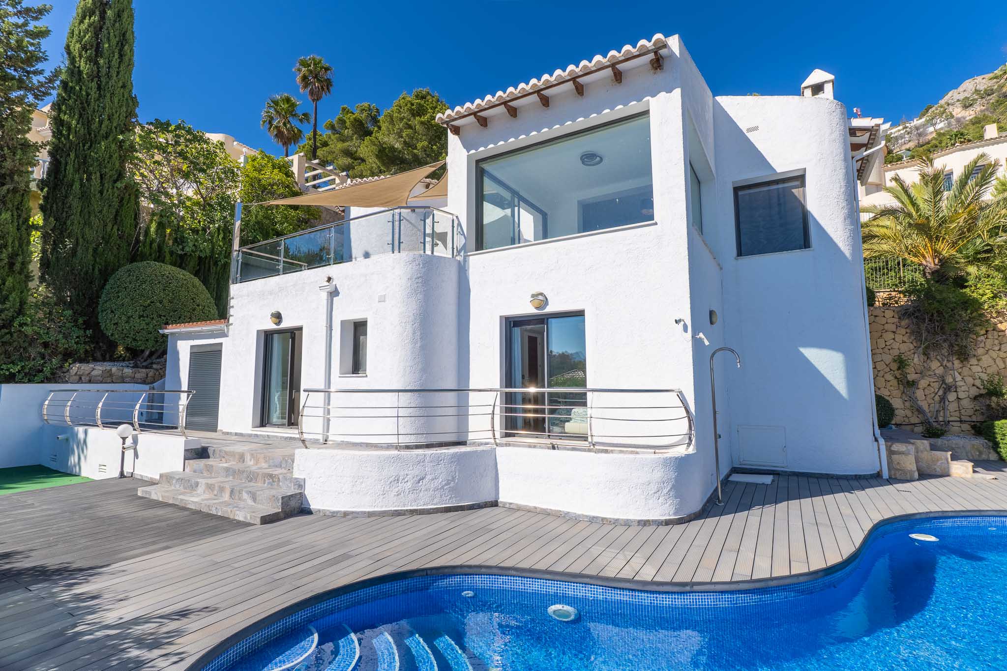 Witte villa-exterieur met grote ramen en terrassen met glazen balustrades. Het blauwe mozaïekzwembad en het grijze composietdek zijn omringd door cipressen en palmbomen.