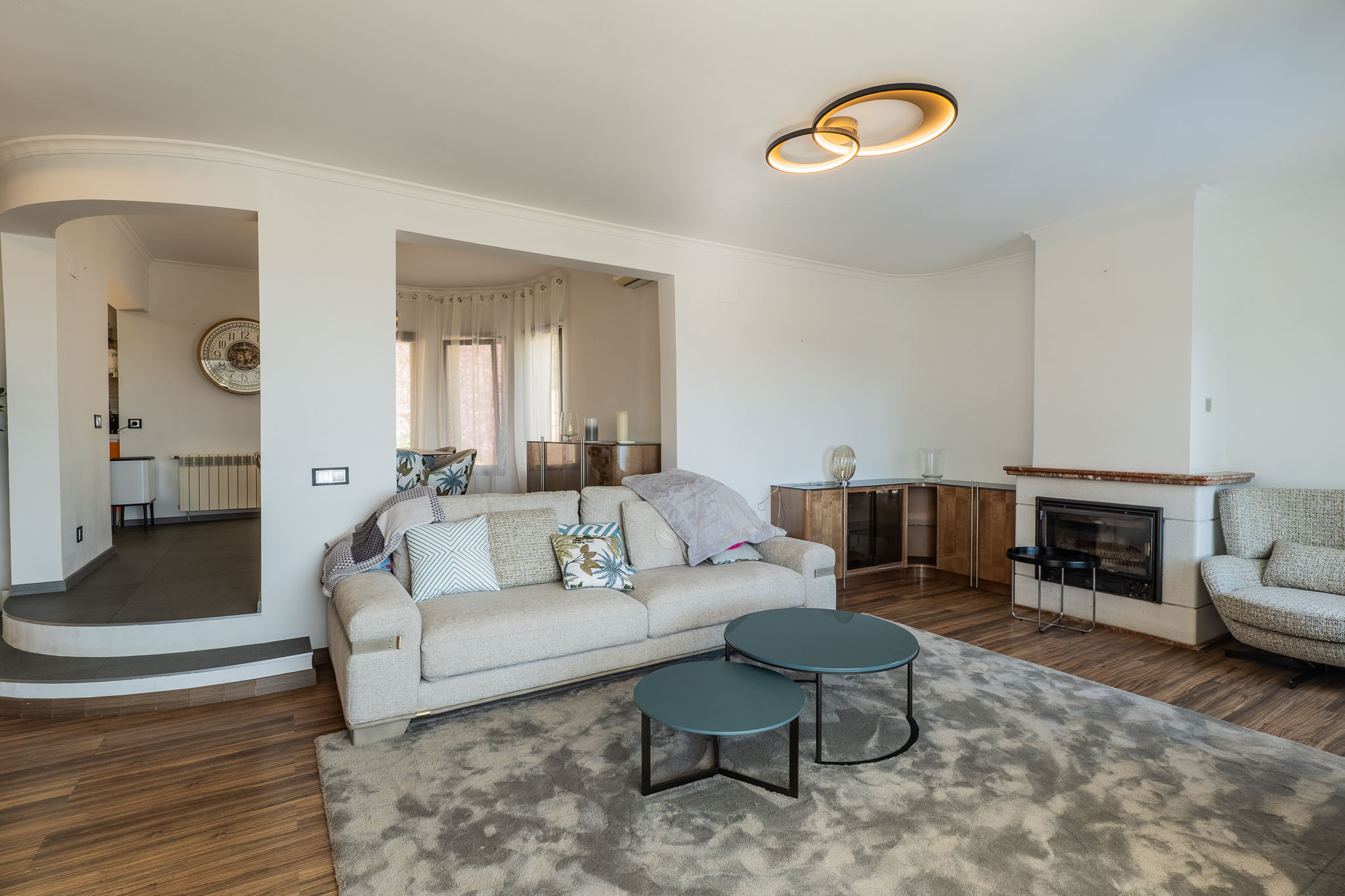 Woonkamer met open haard, roodachtig marmeren schouw en moderne plafondlamp. Een lichte bank en tealkleurige salontafels op een grijs vloerkleed leiden naar een verhoogd gedeelte.