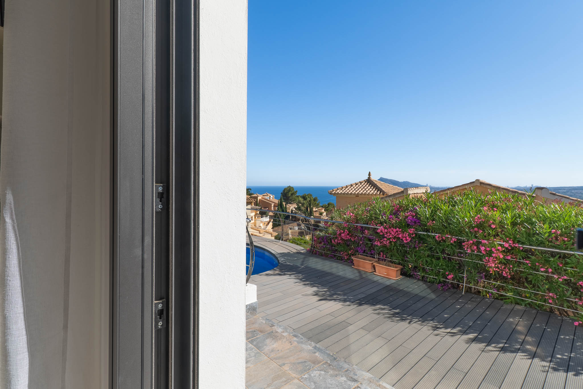 Zicht vanuit de schuifdeur op het terras met composiet vlonder, een glimp van het zwembad en stalen balustrade. De Middellandse Zee en traditionele daken zijn zichtbaar.