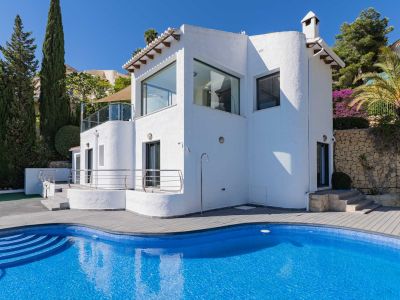 Buitenaanzicht van de villa, met een gebogen blauw mozaïekzwembad, grijze composiet vlonder, buitendouche en mediterrane beplanting.