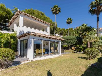 Buitenaanzicht van de villa: witte gevel, traditionele dakpannen, grote glazen puien. De vlakke tuin biedt een gazon en een terras met pergola, omgeven door palmen en struiken.