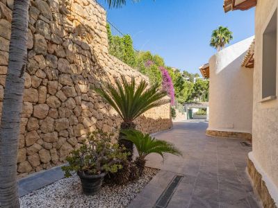 Buitenpad met robuuste natuurstenen keermuur en mediterrane beplanting, grenzend aan een gebogen witte stucgevel in fel zonlicht.