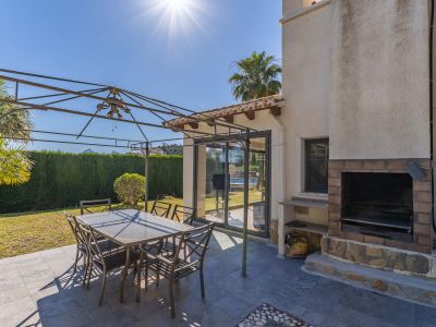 Buitenpatio met donkergrijze tegels, een metalen pergola boven een eettafel en een ingebouwde bakstenen barbecue. Glazen schuifdeuren bieden toegang tot binnen.
