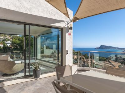 Buitenterras met eettafel en stoelen, deels overschaduwd. Het biedt panoramisch zee- en kustzicht via een glazen balustrade, en verbindt met een lichte binnenruimte.