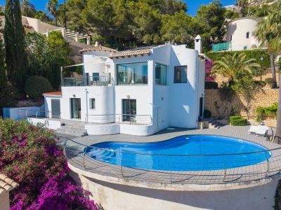 Exterieur van de witte villa met een golvend zwembad en houten terras. Het boventerras met zonnescherm en de levendige bougainvillea versterken de mediterrane sfeer.